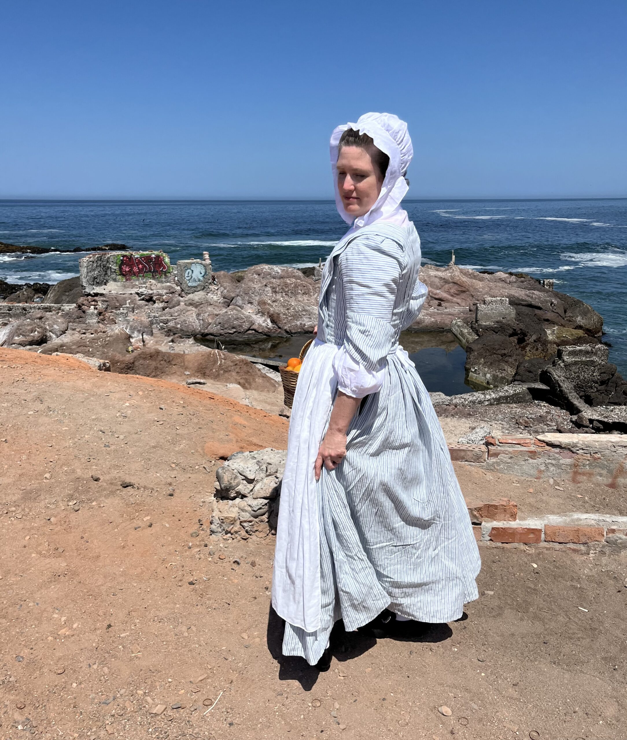 Tabubilgirl looks over her shoulder at the camera. She is wearing a striped linen english nightgown and a white linen lappet cap.