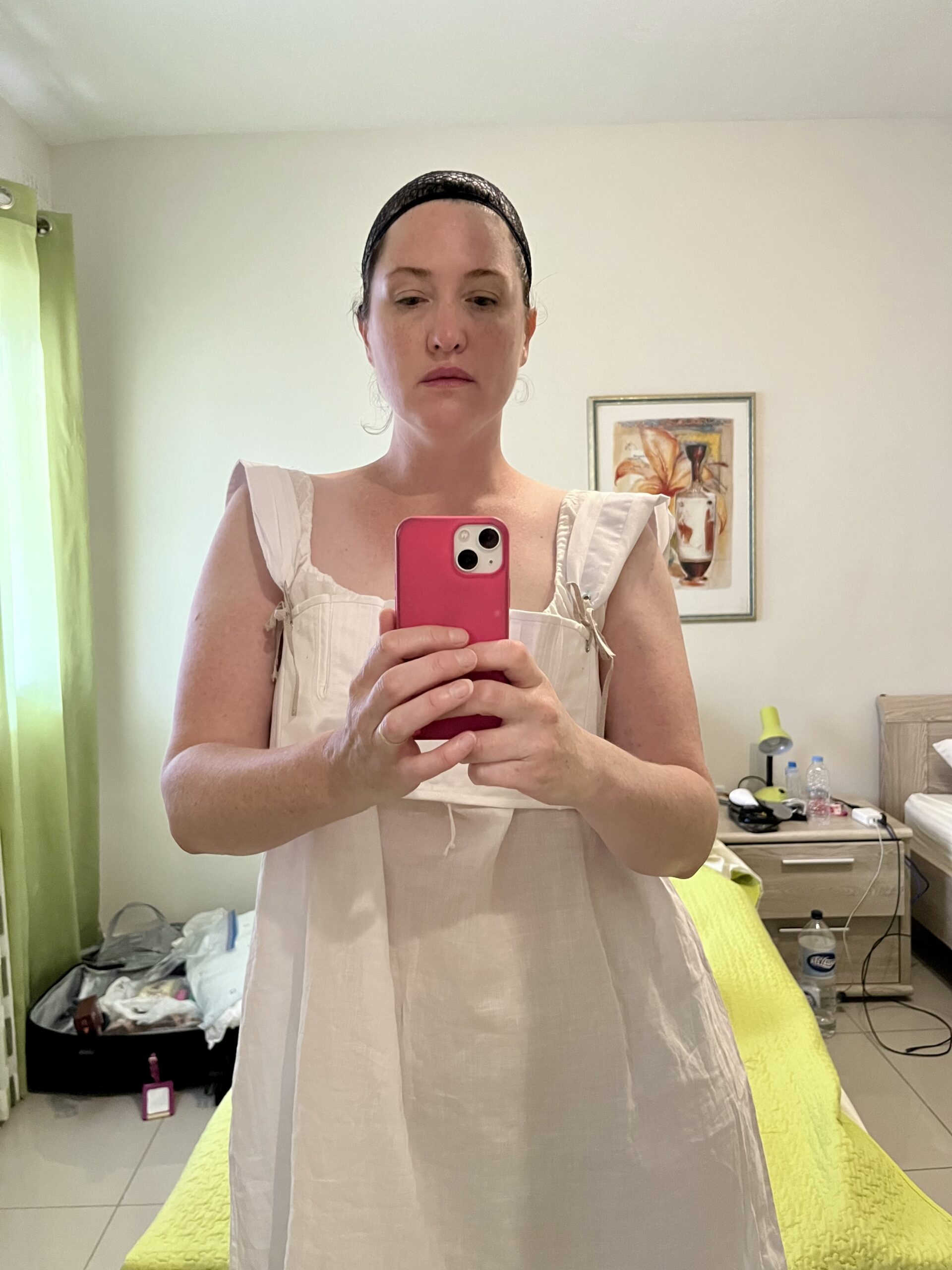 Tabubil girl takes a selfie photo. She is wearing a white cotton shift and a pair of short regency stays. On her head is a wig cap.