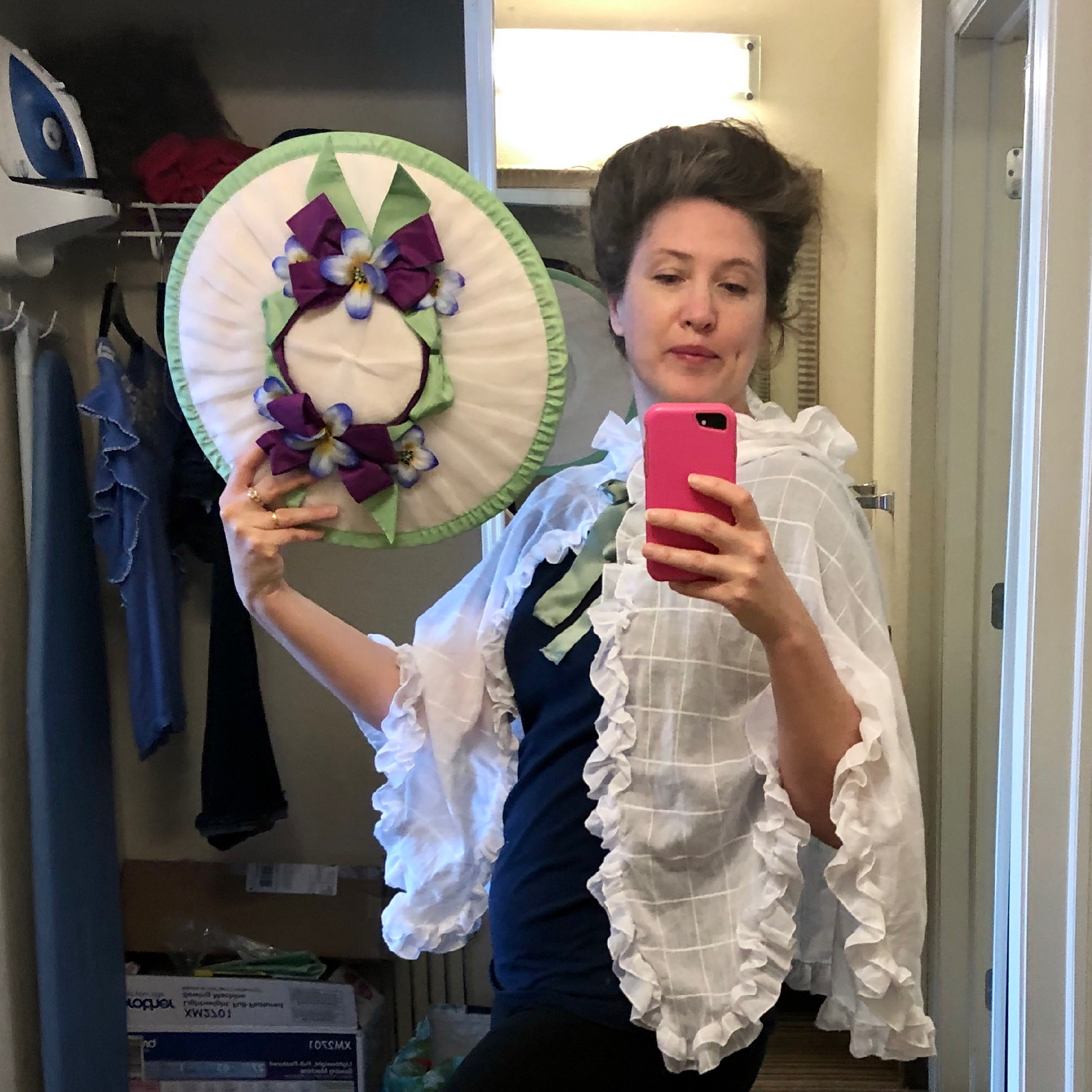 A woman faces the camera holding a cell phone for a mirror selfie. She is wearing a black t-shirt and a white cotton mantelet. In her phone-free hand she is holding a white bergere hat with green ribbon trim and purple flowers