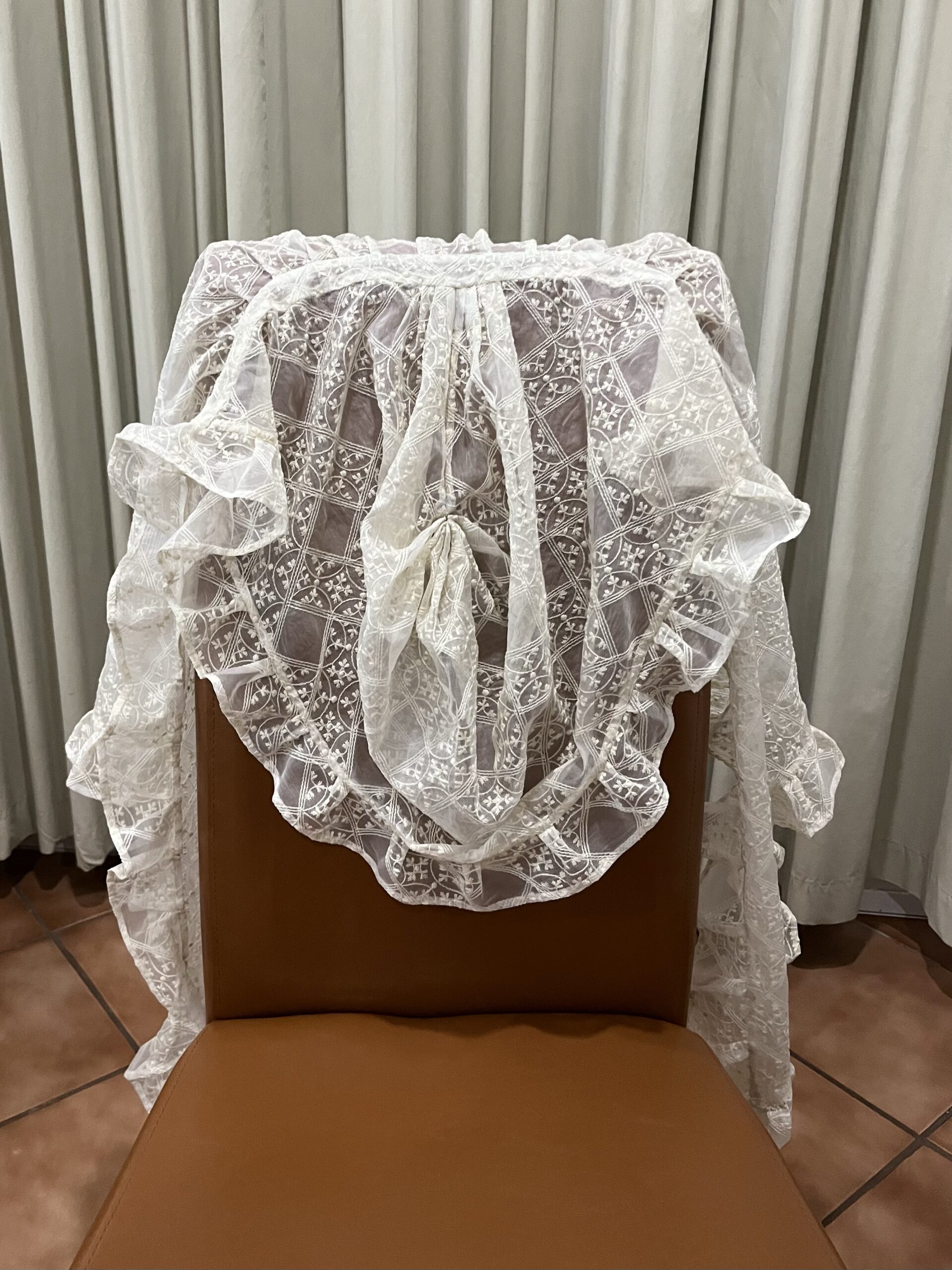 A gauze mantelet lies draped over the back of a chair. The ruffled hood hangs limply.
