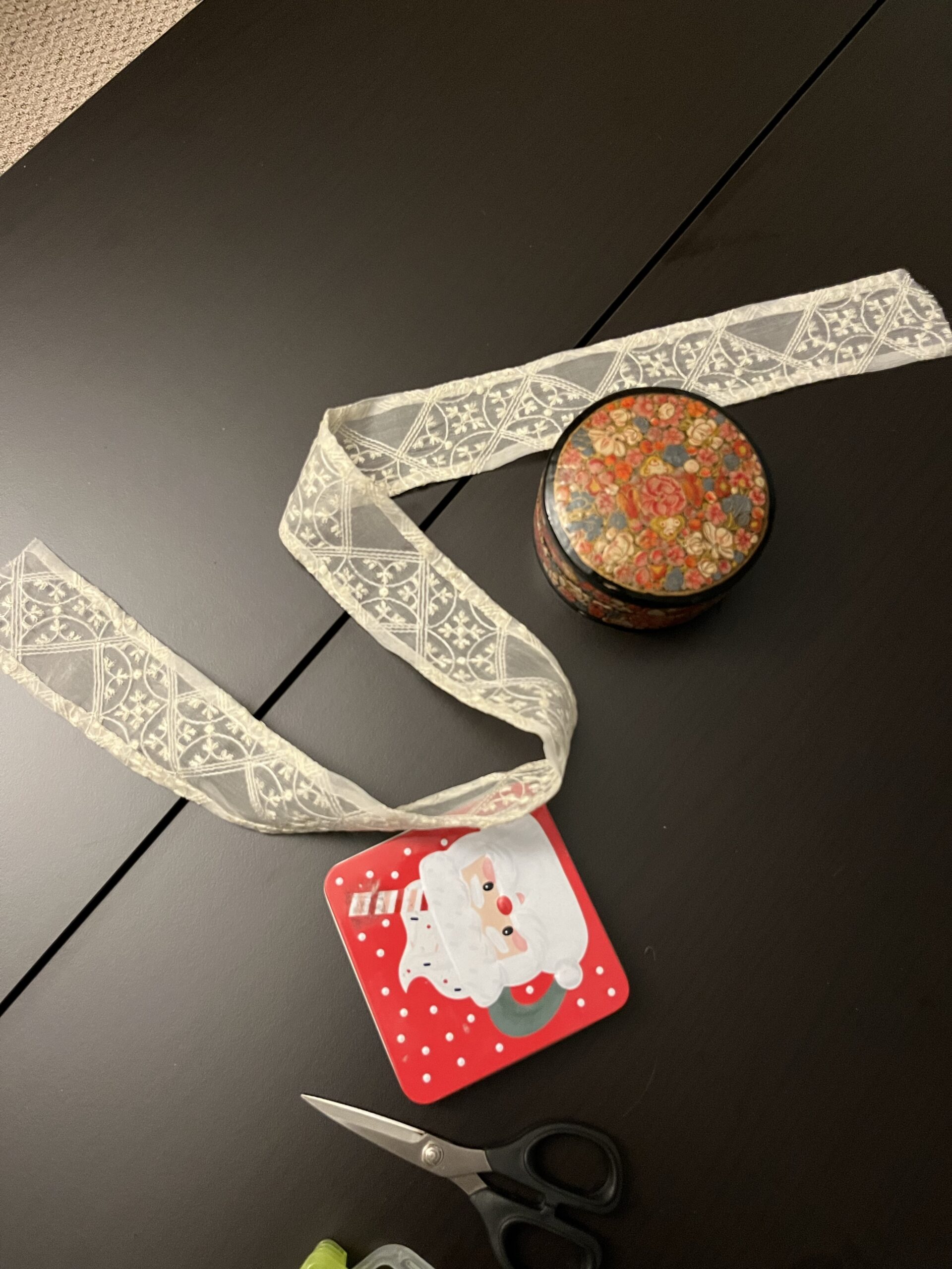 A length of hemmed silk trim lies on a table next to a pair of scissors and a tin with santa's face on it