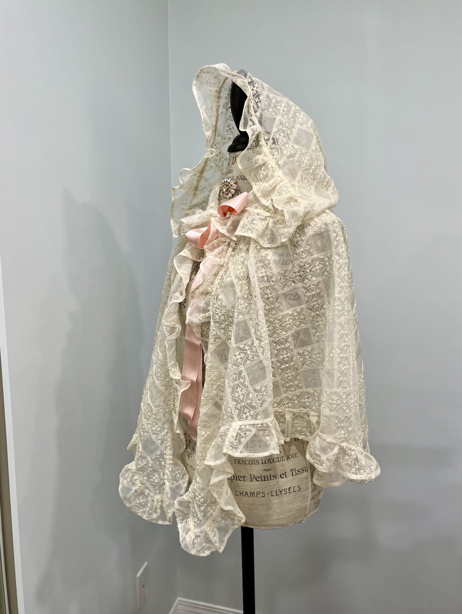 A 3/4 view of a blonde silk gauze mantelet, tied at the neck with a pink silk satin ribbon