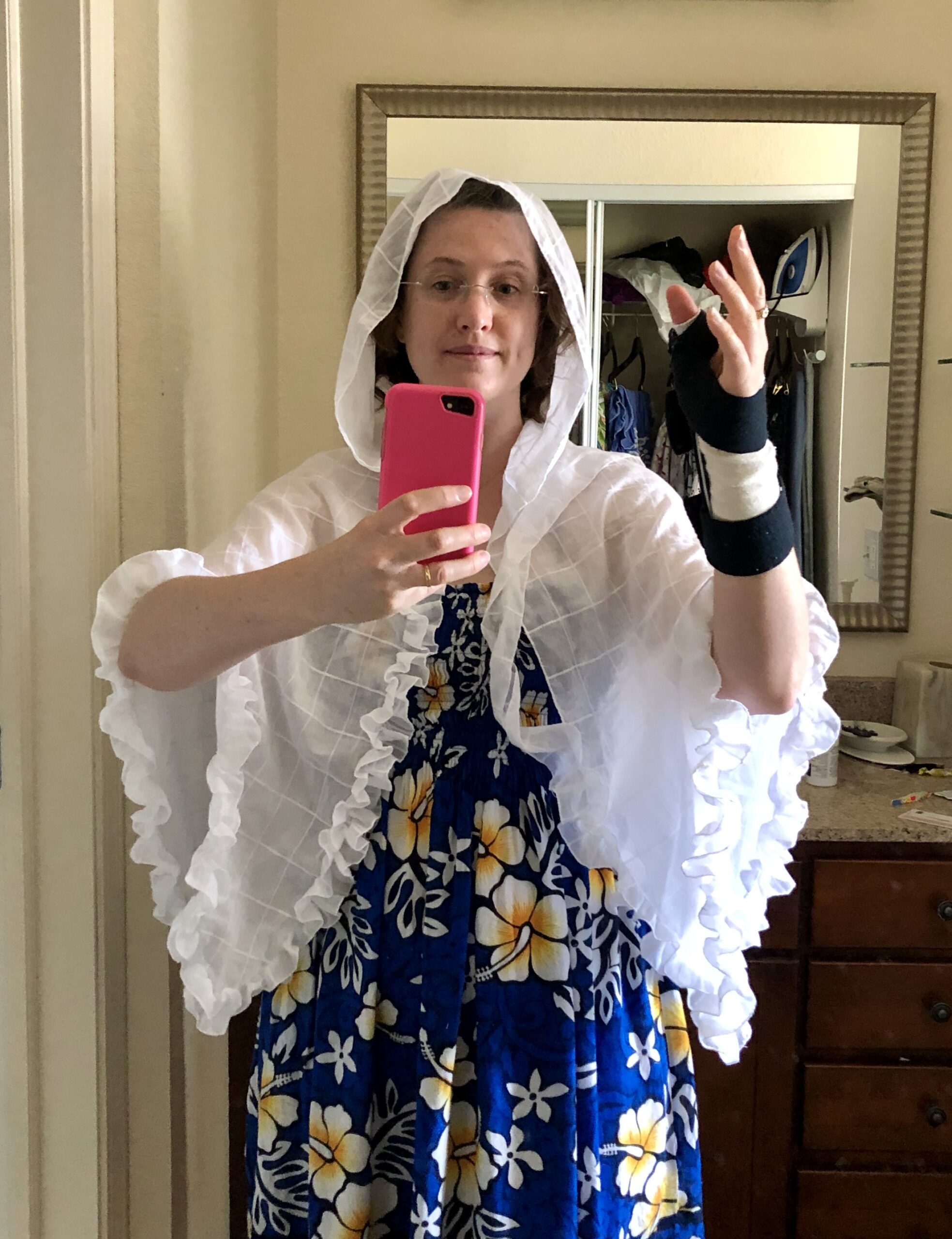 A woman faces the camera holding up a cell phone for a mirror selfie - she is wearing a blue dress with yellow hibiscus flowers and is draped in white cotton mantelet. The ruffle is half attached.
