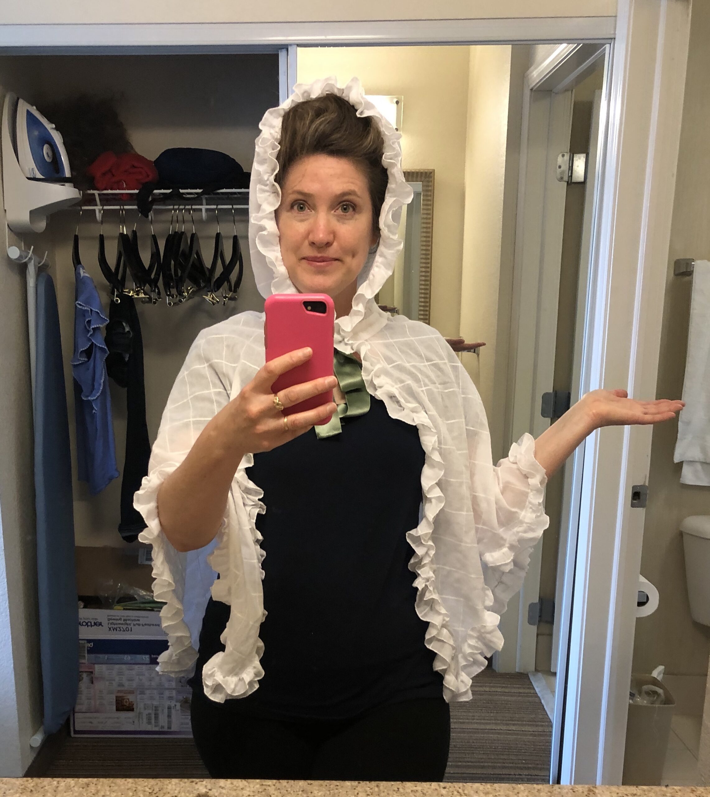 A woman faces the camera holding a cell phone for a mirror selfie. She is wearing a black t-shirt and a white cotton mantelet.