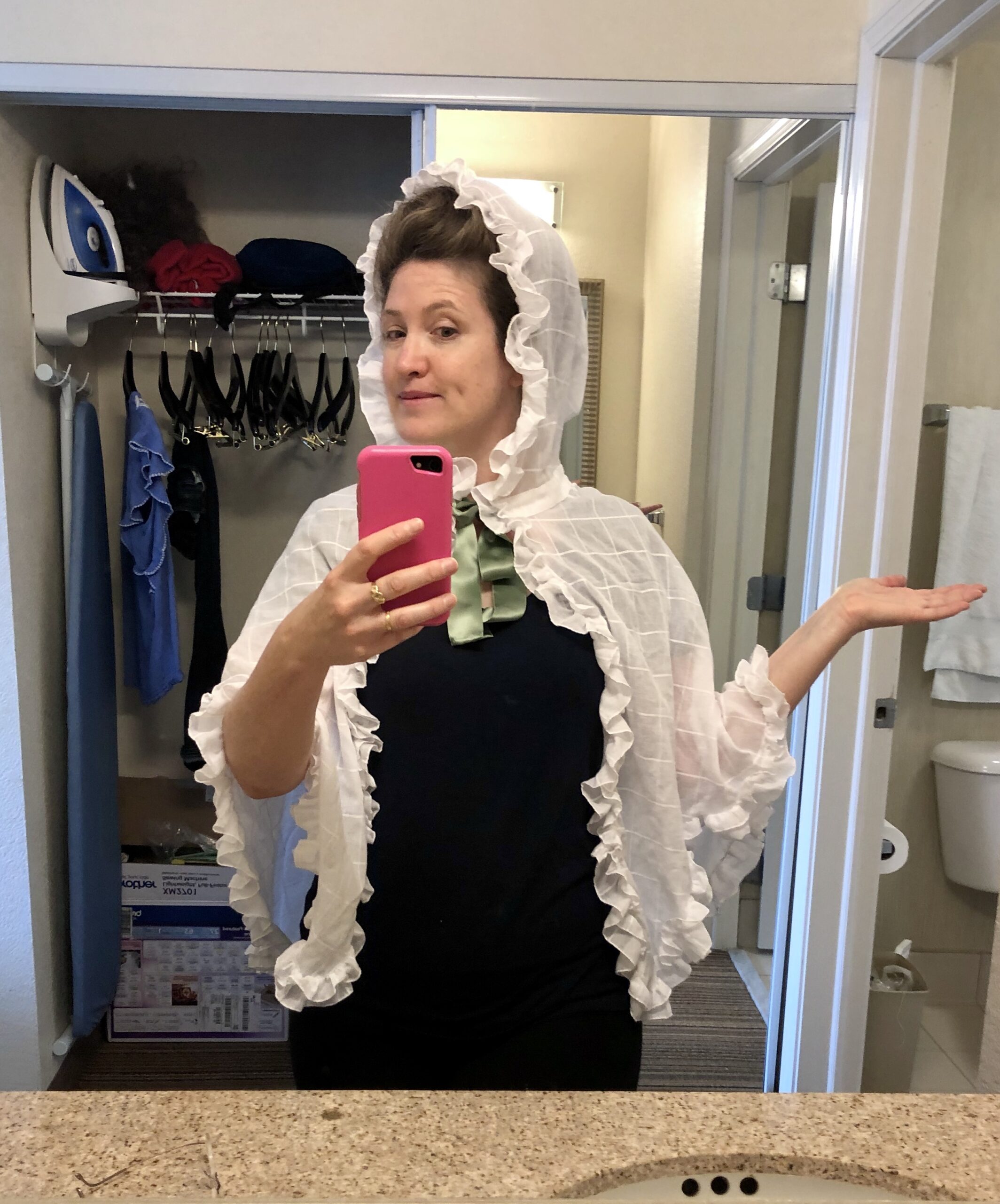A woman stands facing the camera holding a cell phone for a mirror selfie. Her face is slightly turned away She is wearing a black t-shirt and a white cotton mantelet.