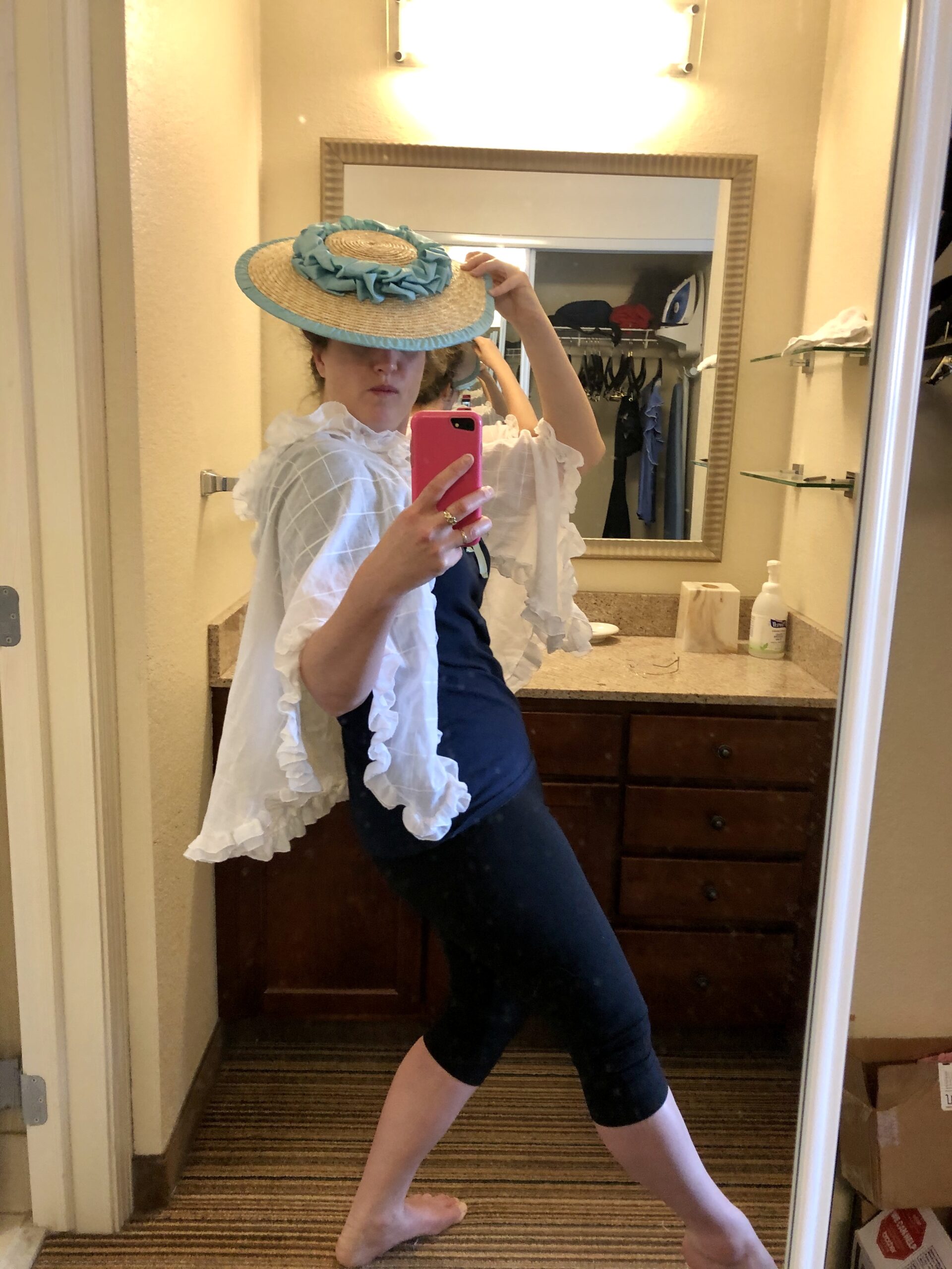 A woman stands for a mirror selfie. She is wearing a white ruffled cotton mantelet and a straw bergere hat trimmed in green ribbon pouffs. She stands in a classic 1950s fashion model broken doll pose