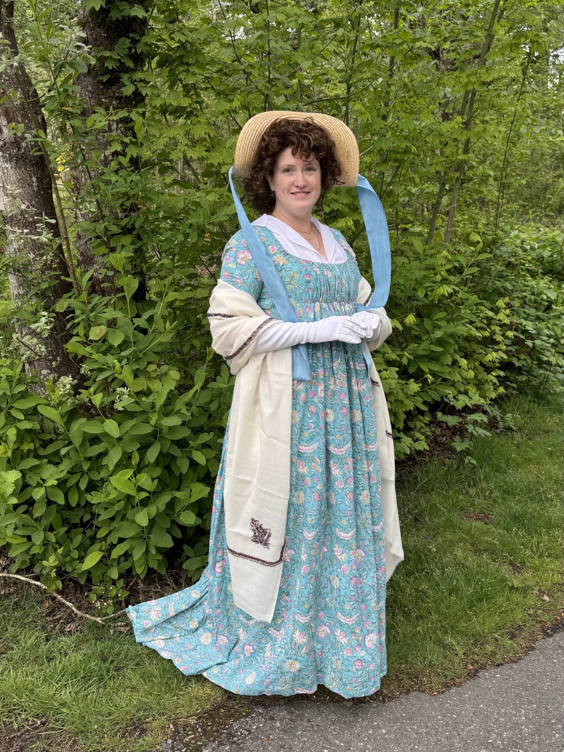 A woman stands at a 3/4 angle to the camera. She is dressed for a spring picnic, in a green floral 1790s round gown and white gloves. There is a long cream wool shawl over her arms. On her head is a large straw hat with blue ribbons flying from it.