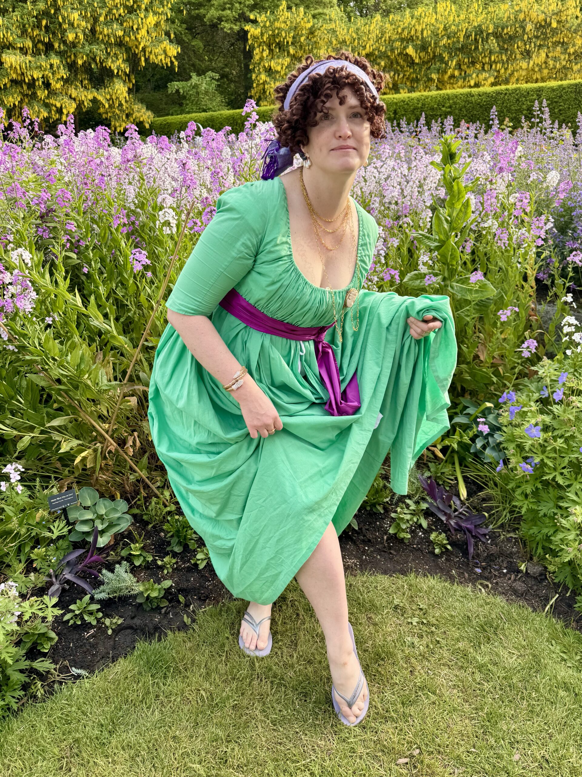 A woman dressed as a high fashion french merveilleuse in a green 1790s gown holds up her skirts to reveal a pair of havaiana sandals