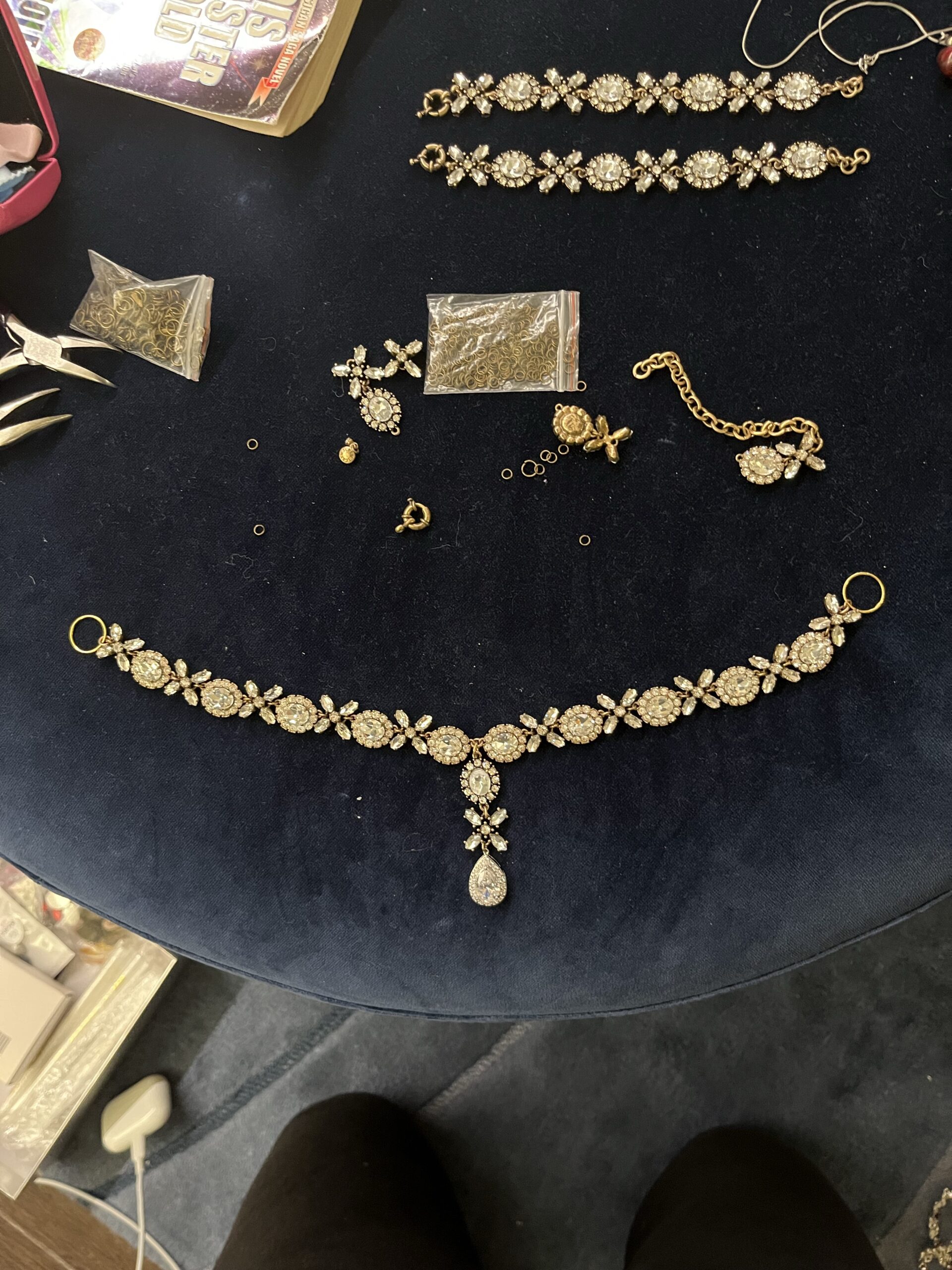 the components of a georgian parure lie on blue velvet ground - a rhinestone necklace and a scatter of jump rings and pliers. then necklace has a pendant drop.