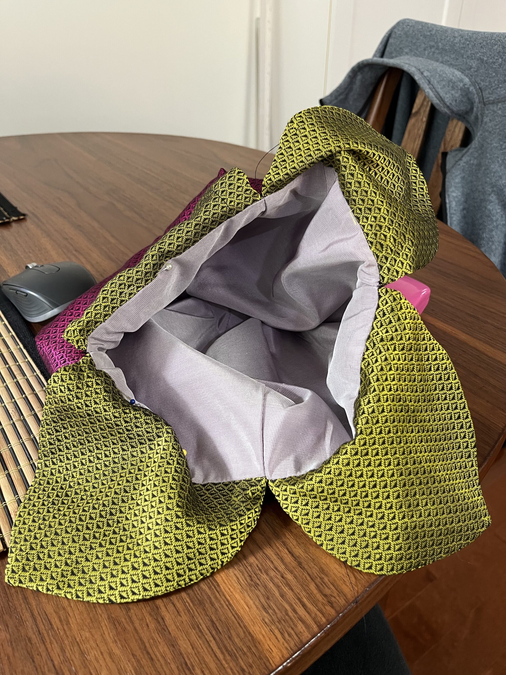 The mouth of a regency reticule gapes open surrounded by green silk leaves. The opening is marked by large pinheads and a needle is stitching into the seam between leaf and lining