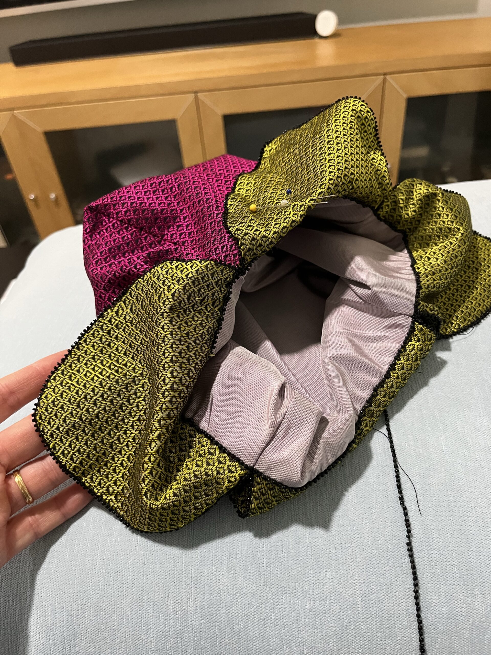 A rasberry regency reticule in pink and green silk lies on a blue sofa. A trim of black glass seed beads is being stitched along the seams.