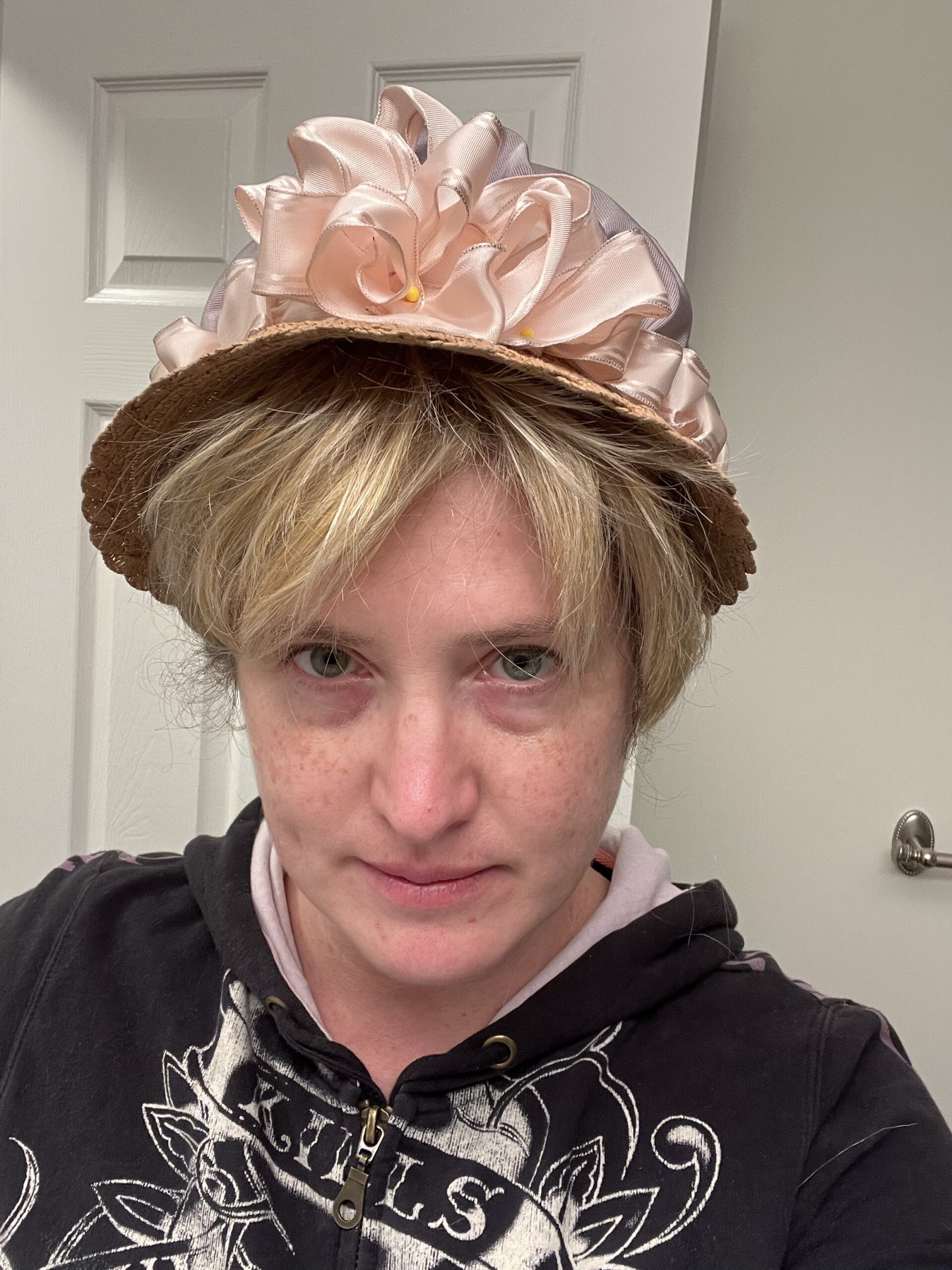 head-shot of a woman wearing a short blonde wig and a straw hat crowned with pink ribbons