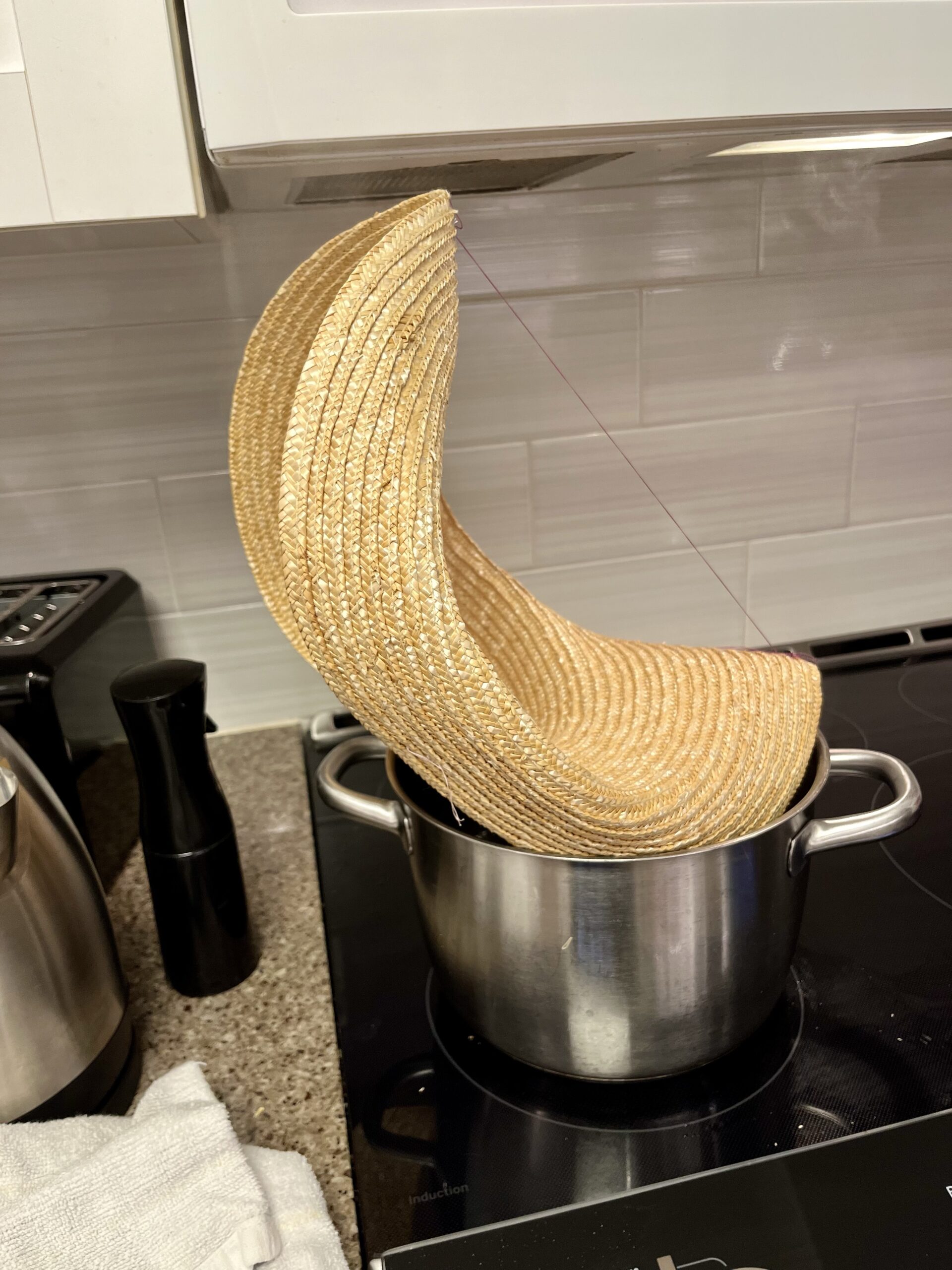 A large straw hat is sitting on top of a pot of water on a stove. The hat is bent into a curve and the bent edges are connected by a taut string