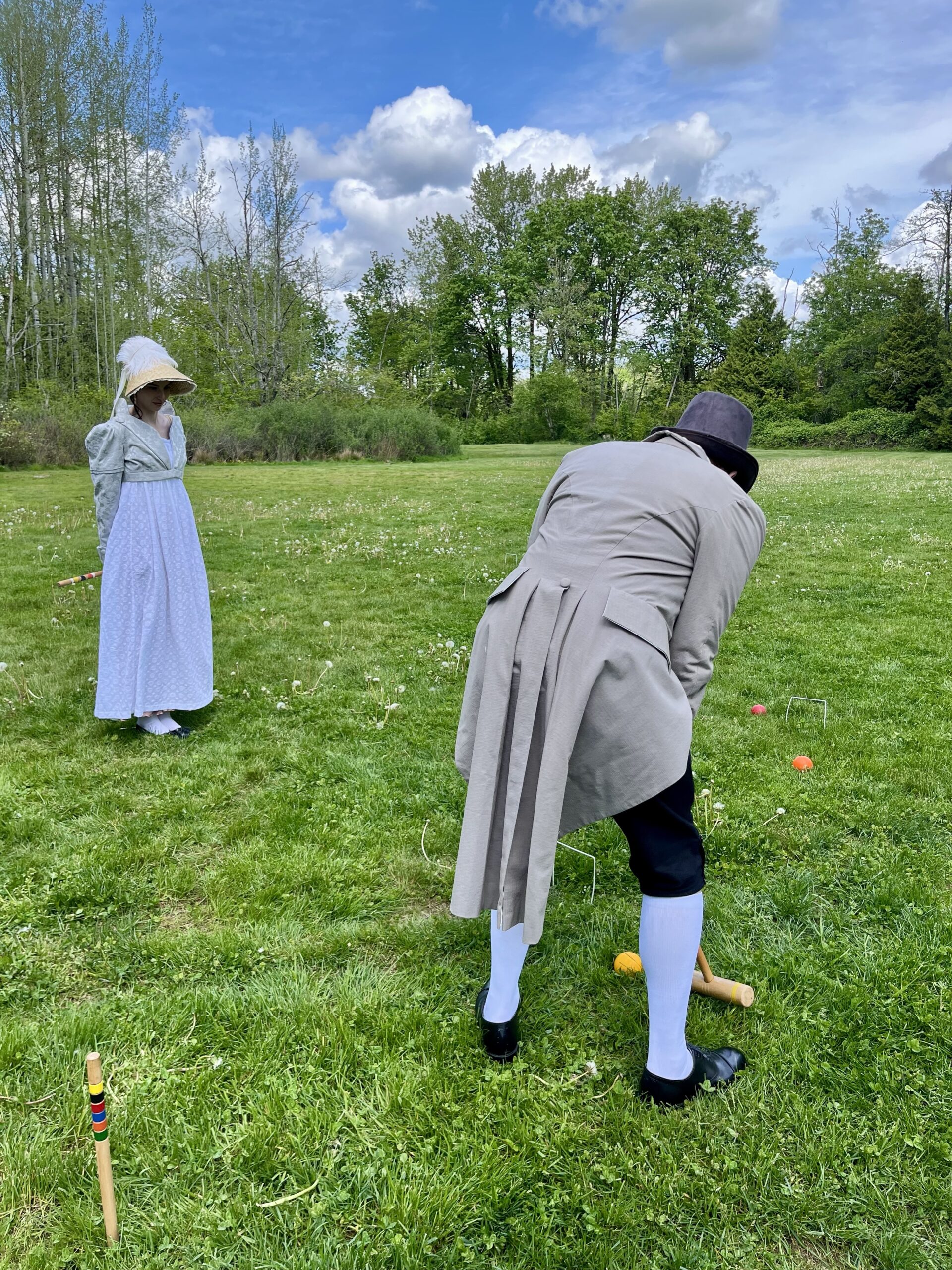 A man in regency breeches, stocking and grey linen coat has his back to the camera. He is crouches over a croquet mallet. In the distance to the left stands a woman in a long regency gown and tall bonnet