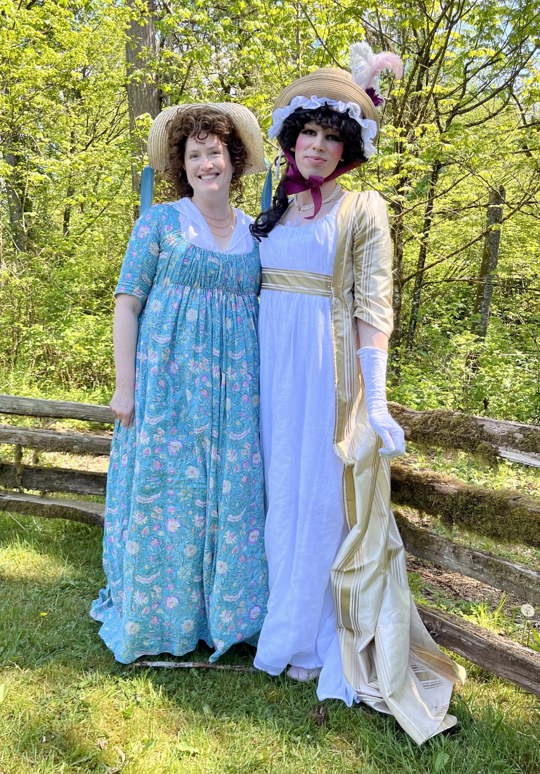Two people face the camera. The person on the left is wearing a green floral 1790s round gown. The person on the right is wearing a white 1790s round gown and a yellow silk striped overgown.