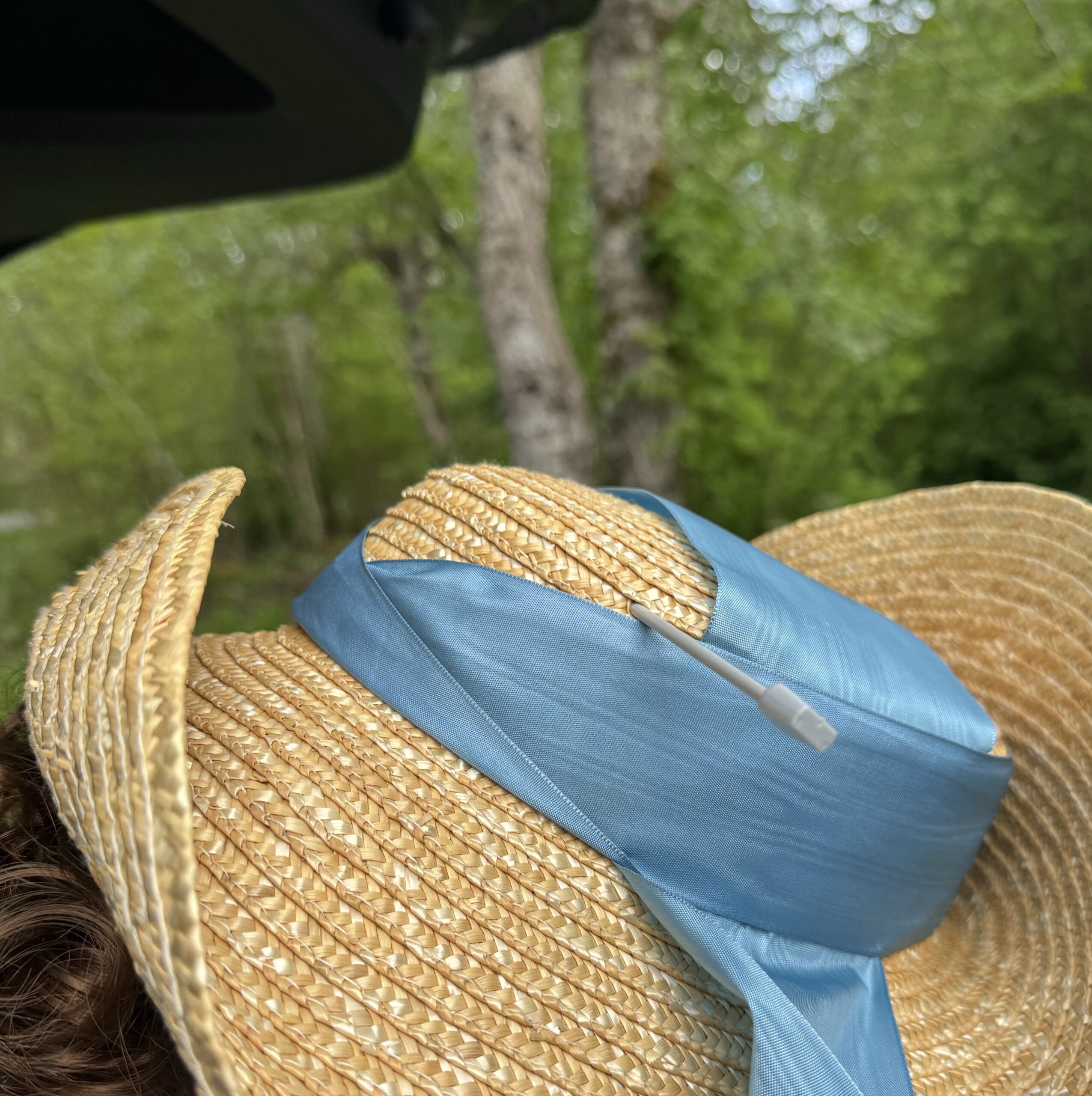 Close up view of a straw hat trimmed with blue moire ribbons and stabbed through the crown with a knitting needle.