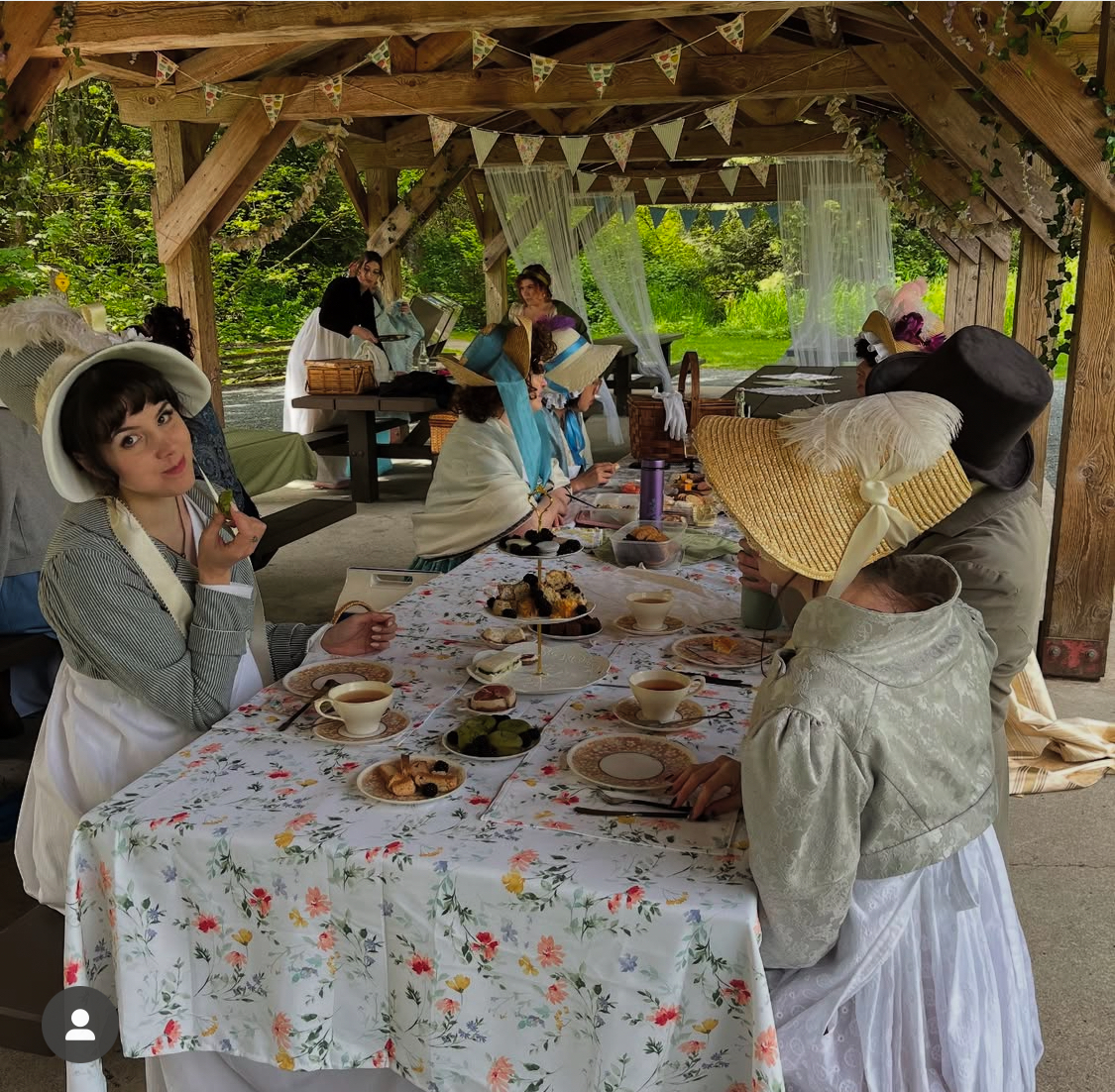 Members of the Hemwick Regency Society sit down for their spring picnic at tables covered in bright floral tablecloths. They are dressed in Regency gowns and bonnets