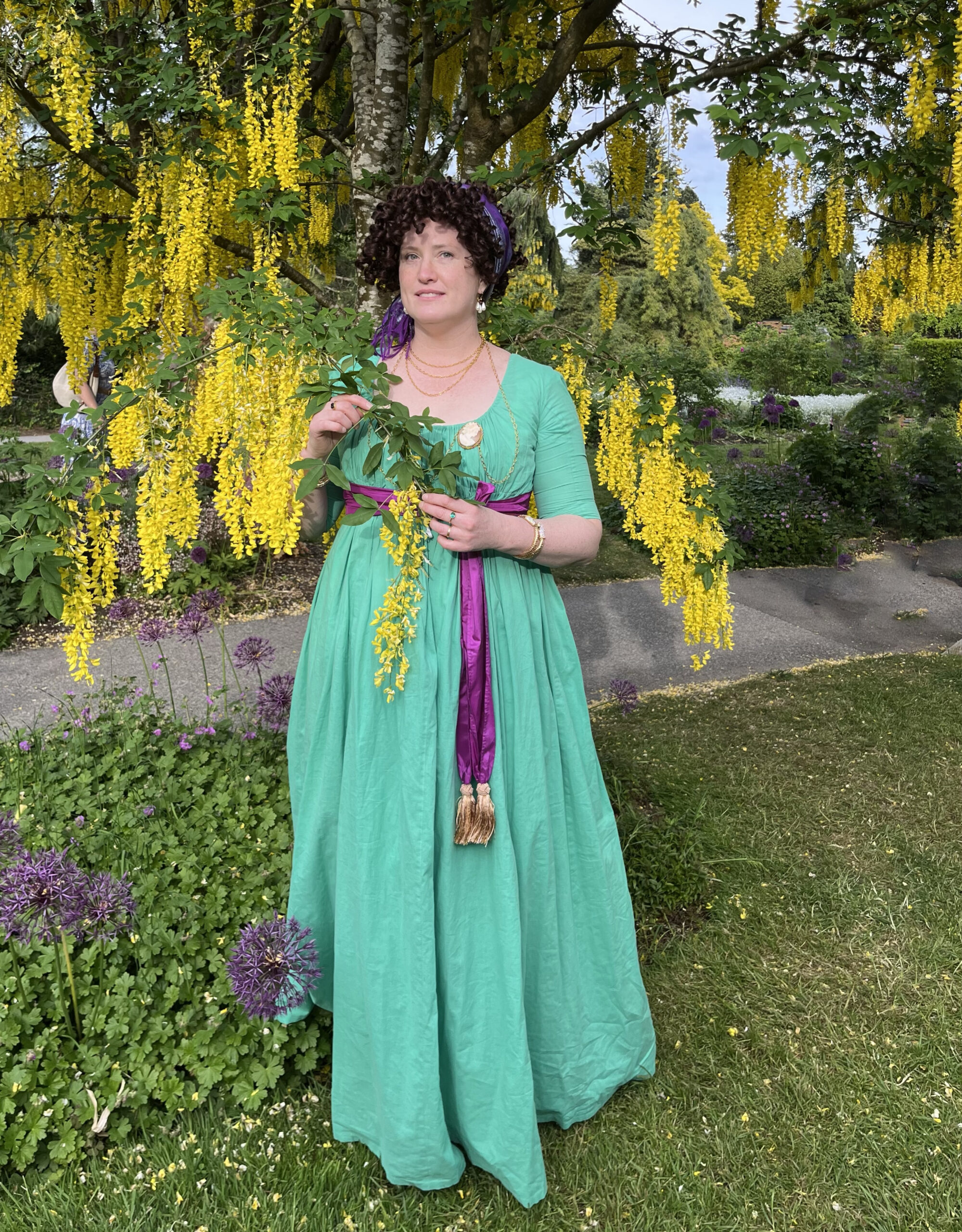 a woman dressed as a high fashion merveilleuse in a green 1790s round gown stands beneath golden laburnum