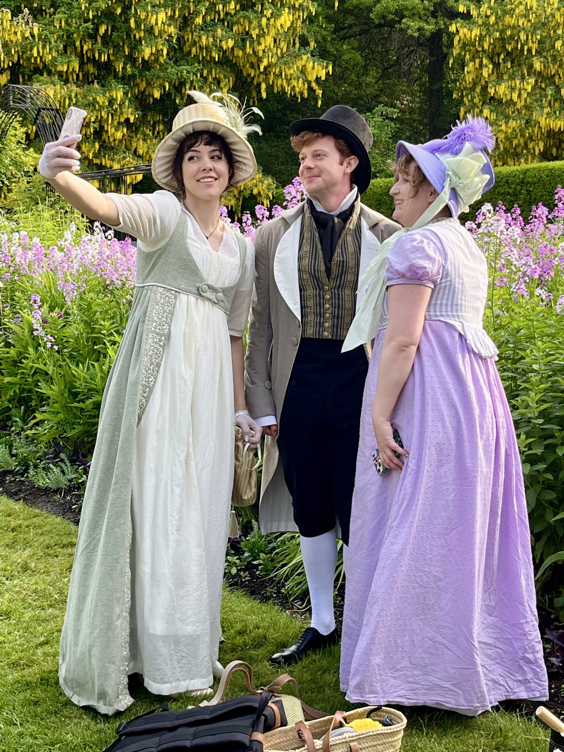 three people in regency clothing pose for a selfie in a garden.