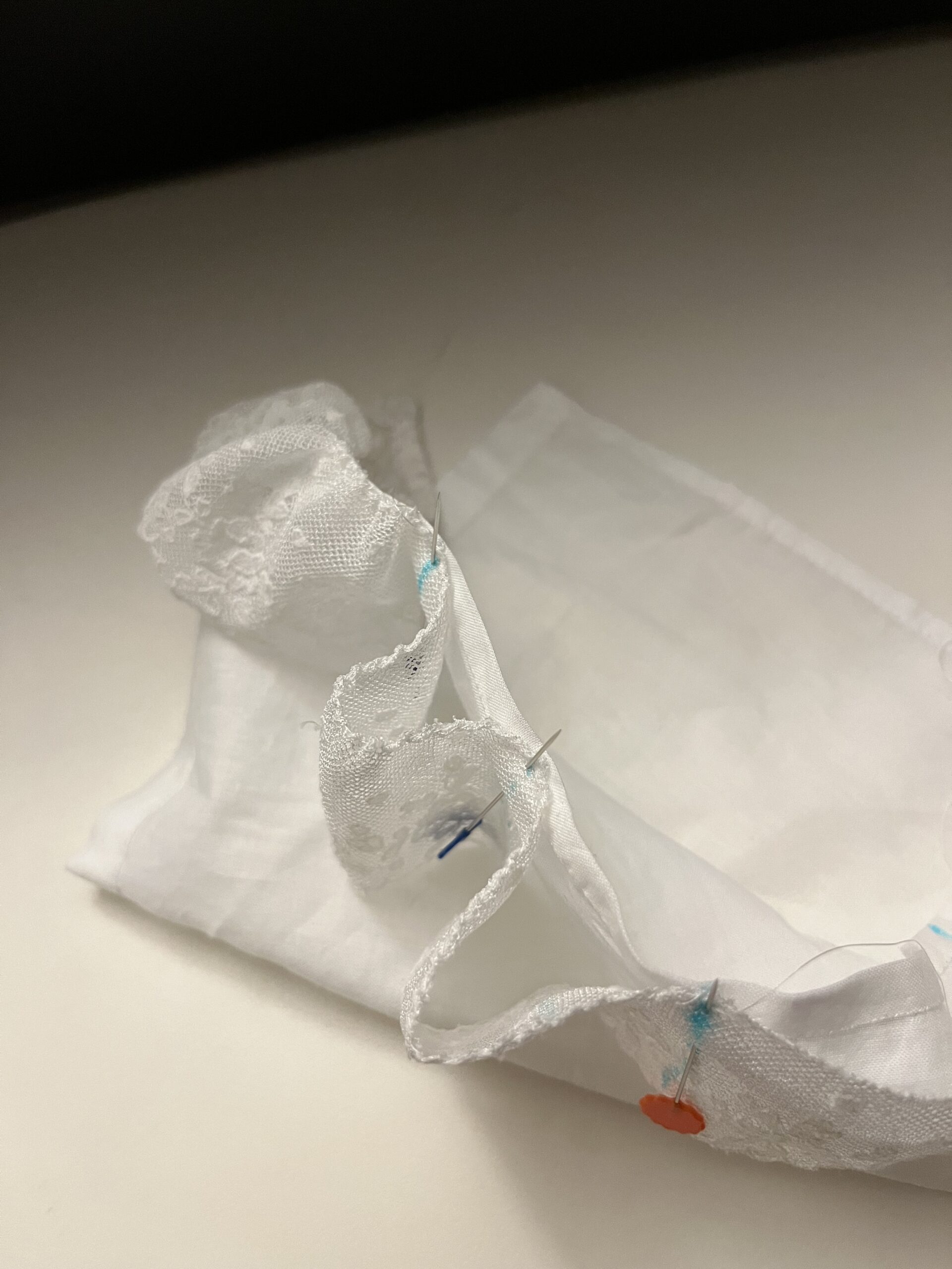 A white piece of fabric with lace pinned to it at intervals. The lace stands up from the fabric in loops