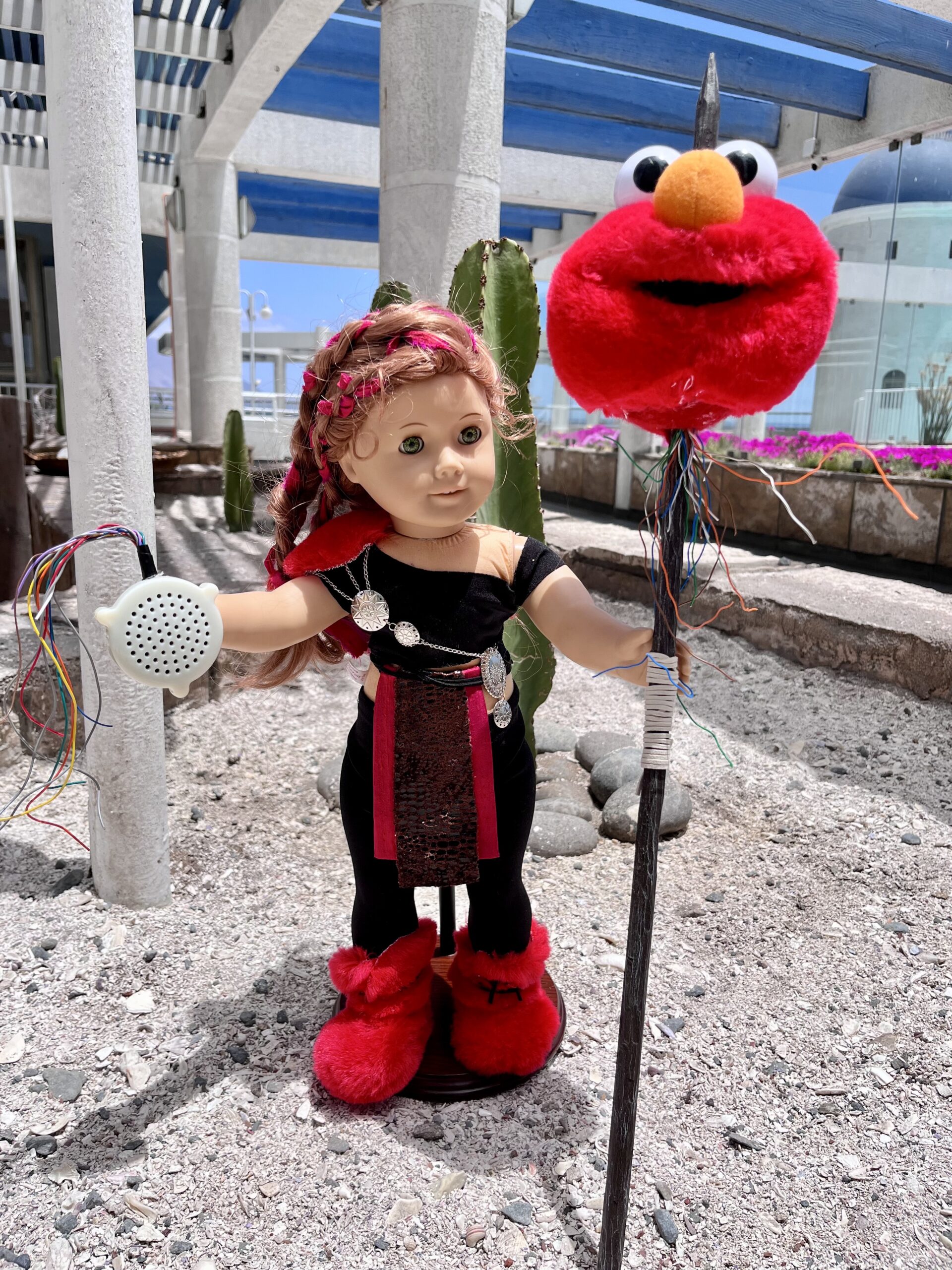 Elmoy, the American Girl Doll Bounty Hunter stands on a sea of white crushed shells. She grasps a pike with the severed head of a tickle me elmo doll