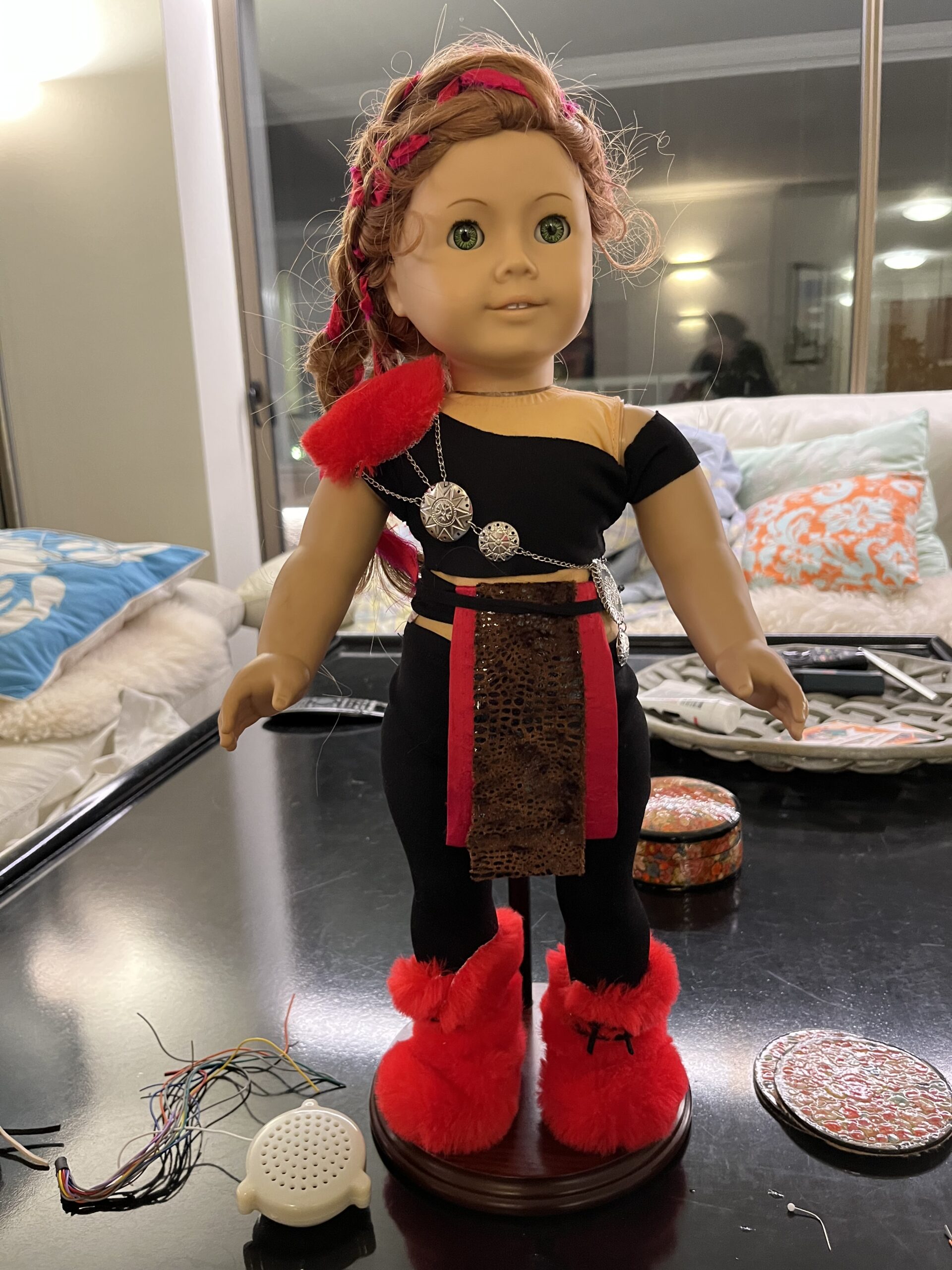 An American Girl doll gets dressed as a bounty hunter. She is standing on a coffee table dressed in black jersey pants and trousers, red fur boots and a red silk tabard