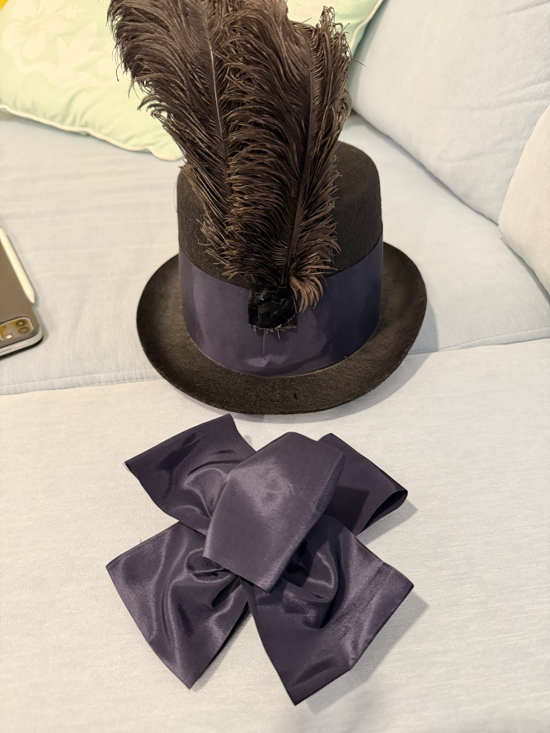 A black hat sits on a sofa. The hat is decorated with a wide ribbon and a spray of ostrich feathers. In front of the hat sits a navy blue bow