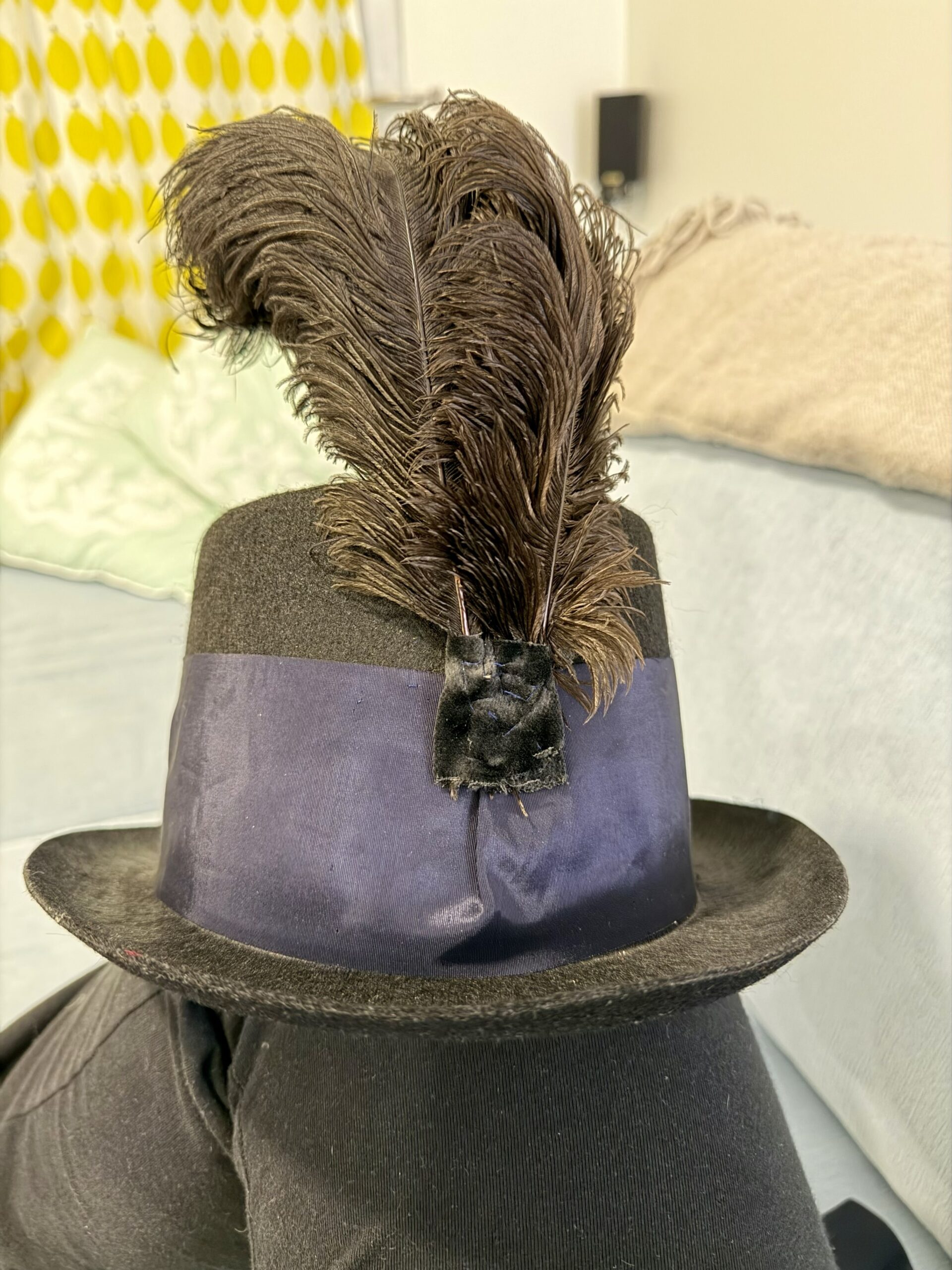 A hat balances on a knee. The hat is decorated with a wide blue ribbon and a spray of black feathers