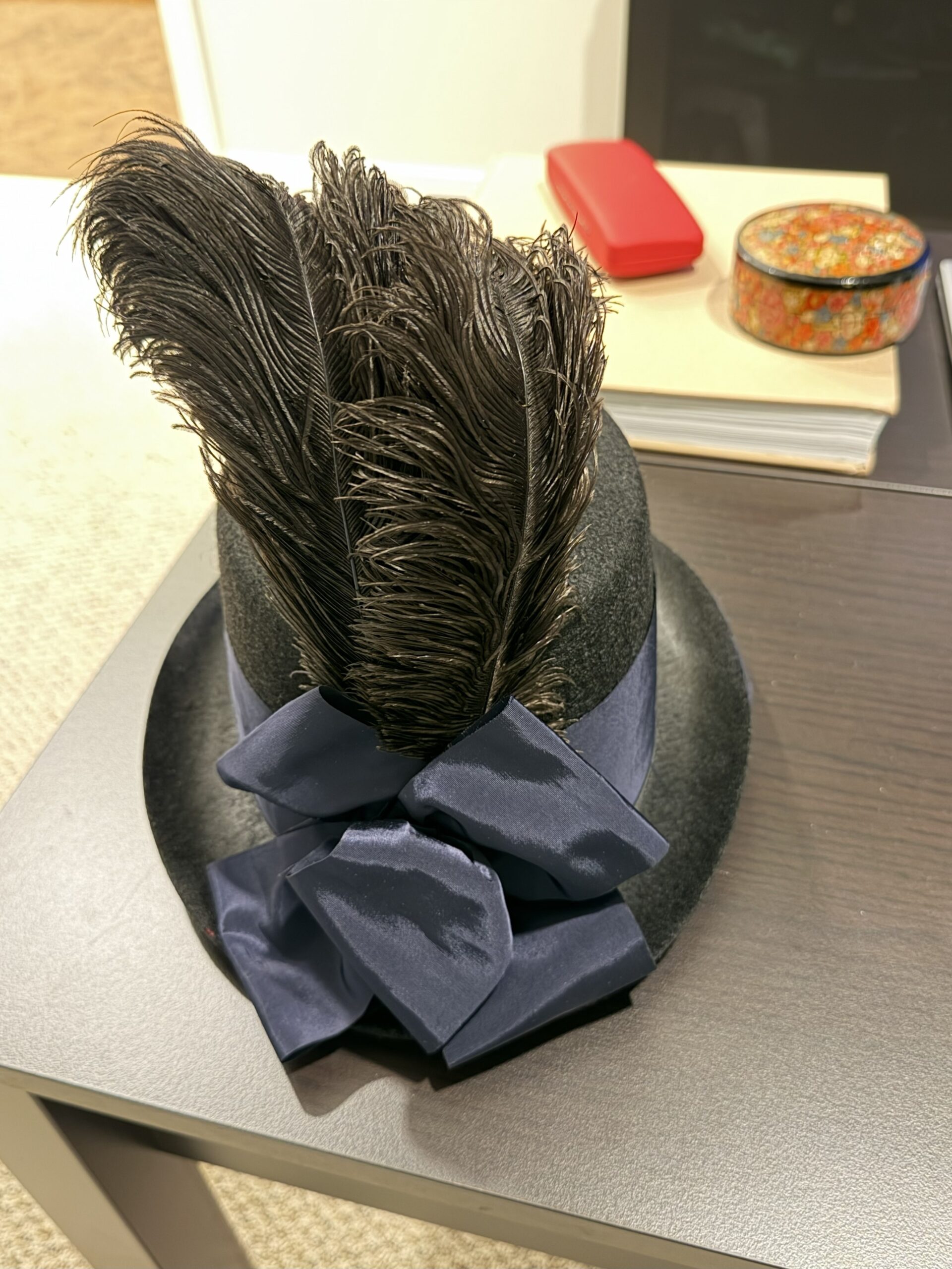 A black regency riding hat sits on a table. it is decorated with a blue bow and a spray of feathers