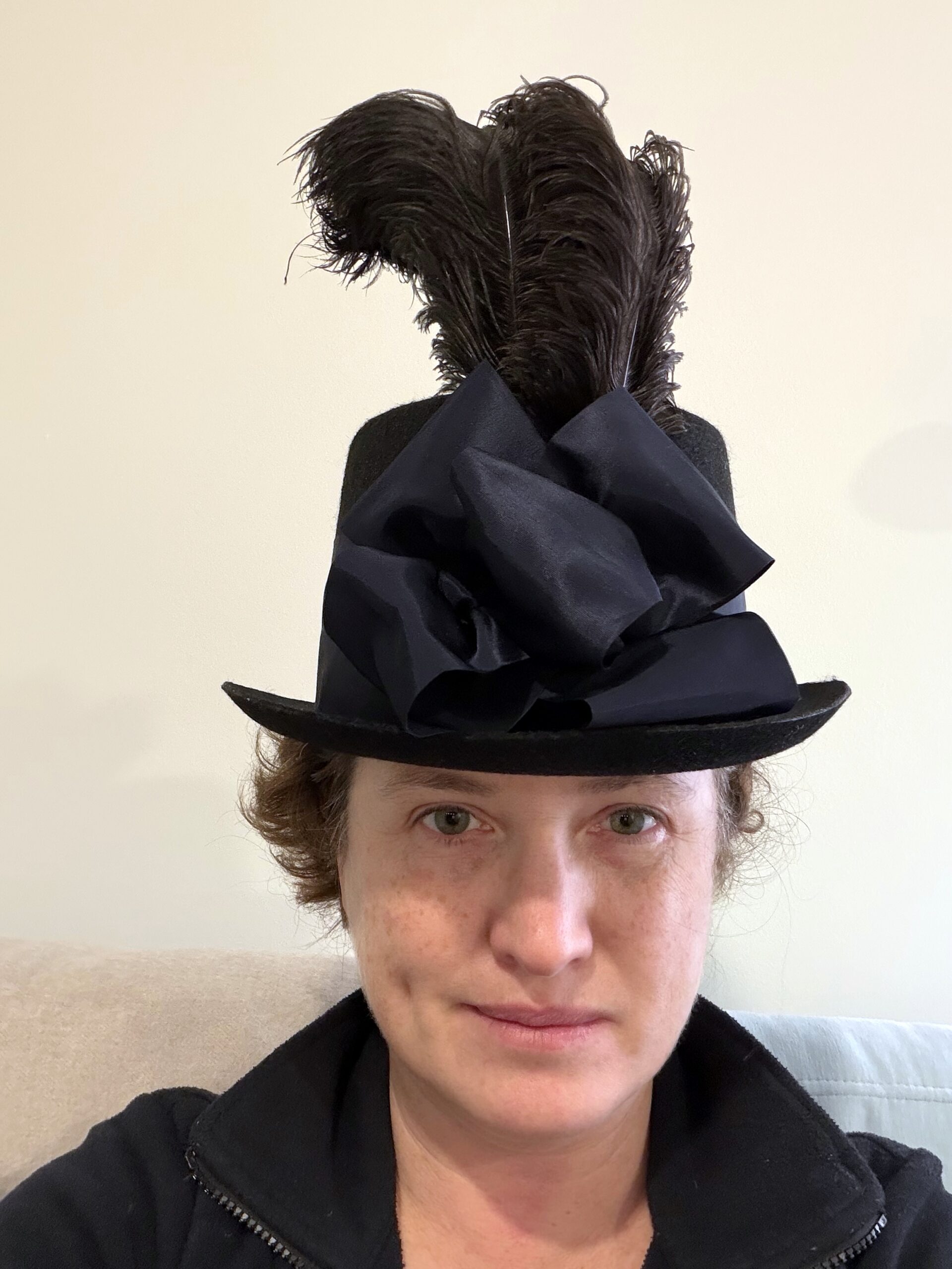 A woman looks at the camera, She is wearing a black regency riding hat with a large bow and two tall black feathers on the front
