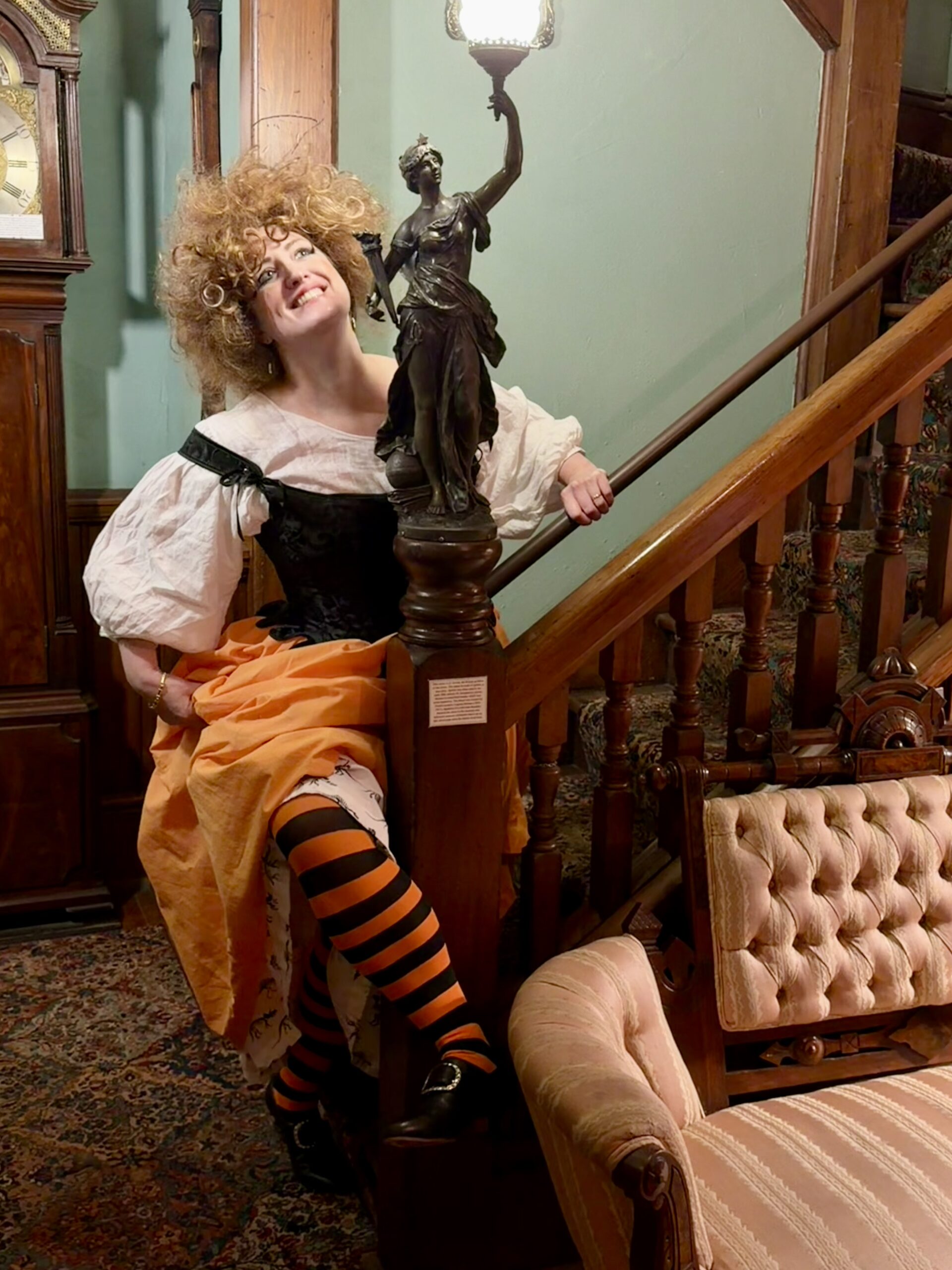 A witchy halloween wench in orange petticoats and orange and black striped stockings smiles around a bronze newel post