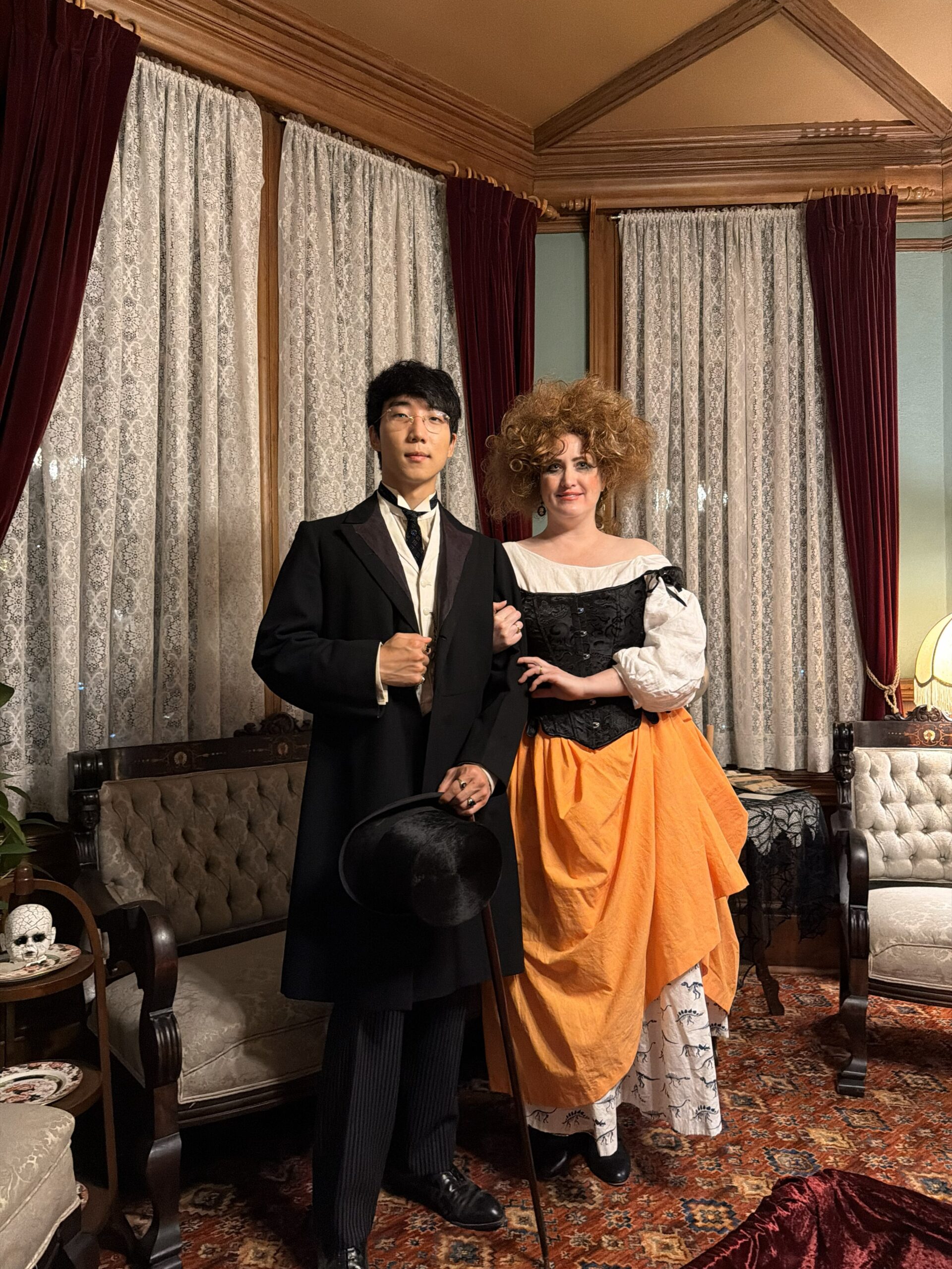 A halloween witch in black corset and orange petticoats is on the arm of a very elegant man in an edwardian suit