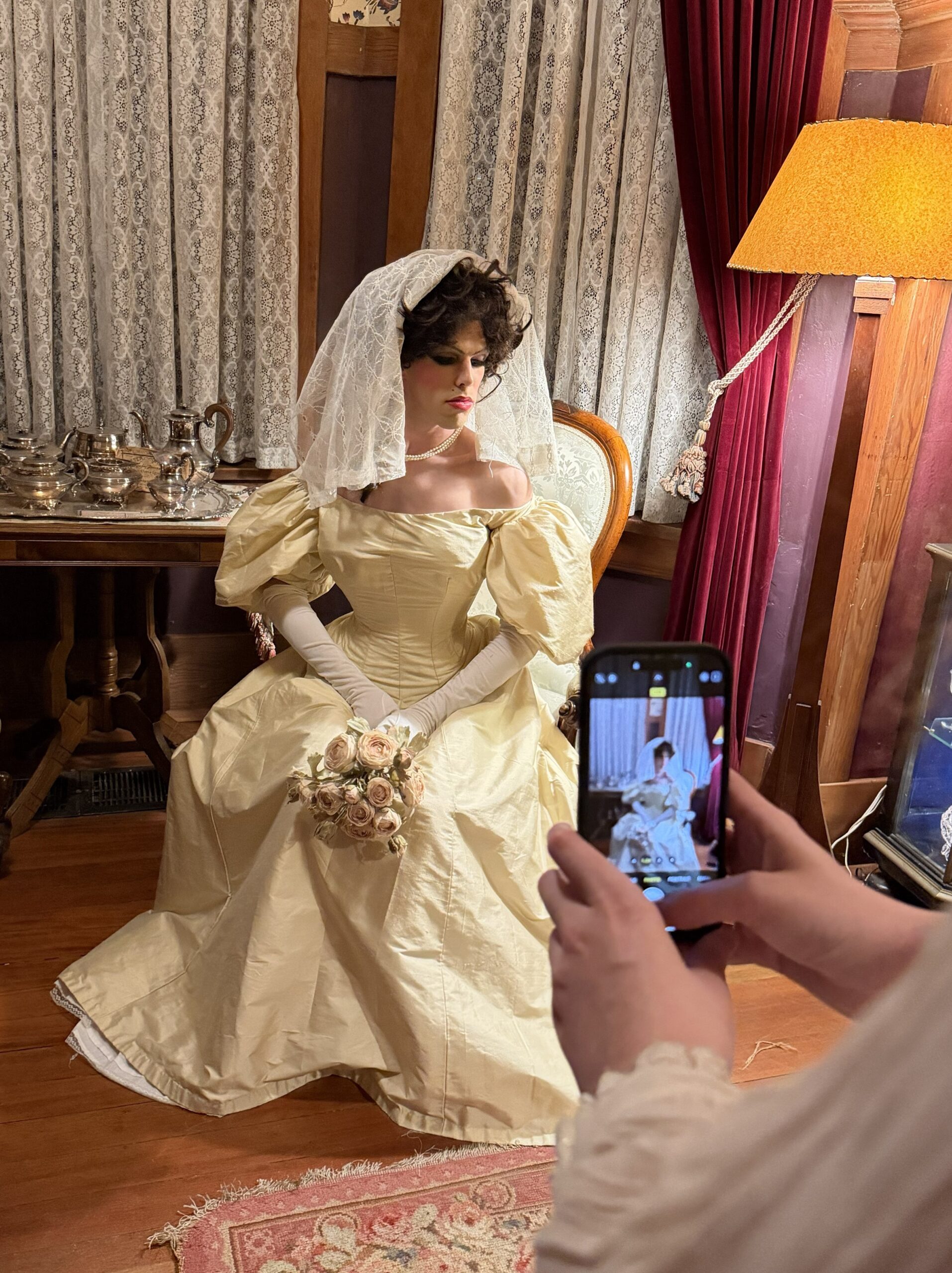 A bride in a white silk edwardian wedding gown and veil sits in a wing chair and looks demurely away from the camera
