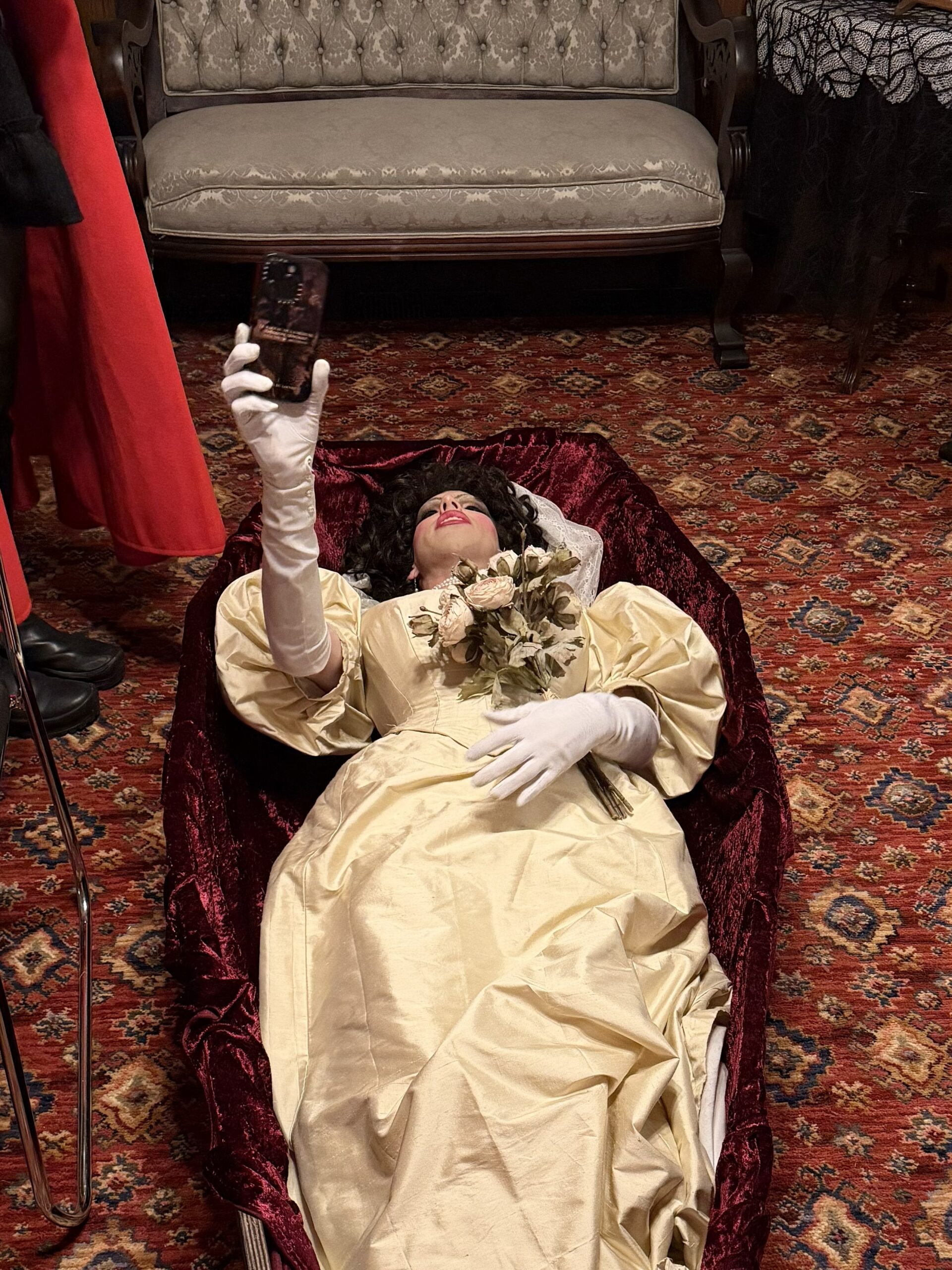 An edwardian bride in a white silk dress lies in a coffin.She's taking a selfie on a cell phone.