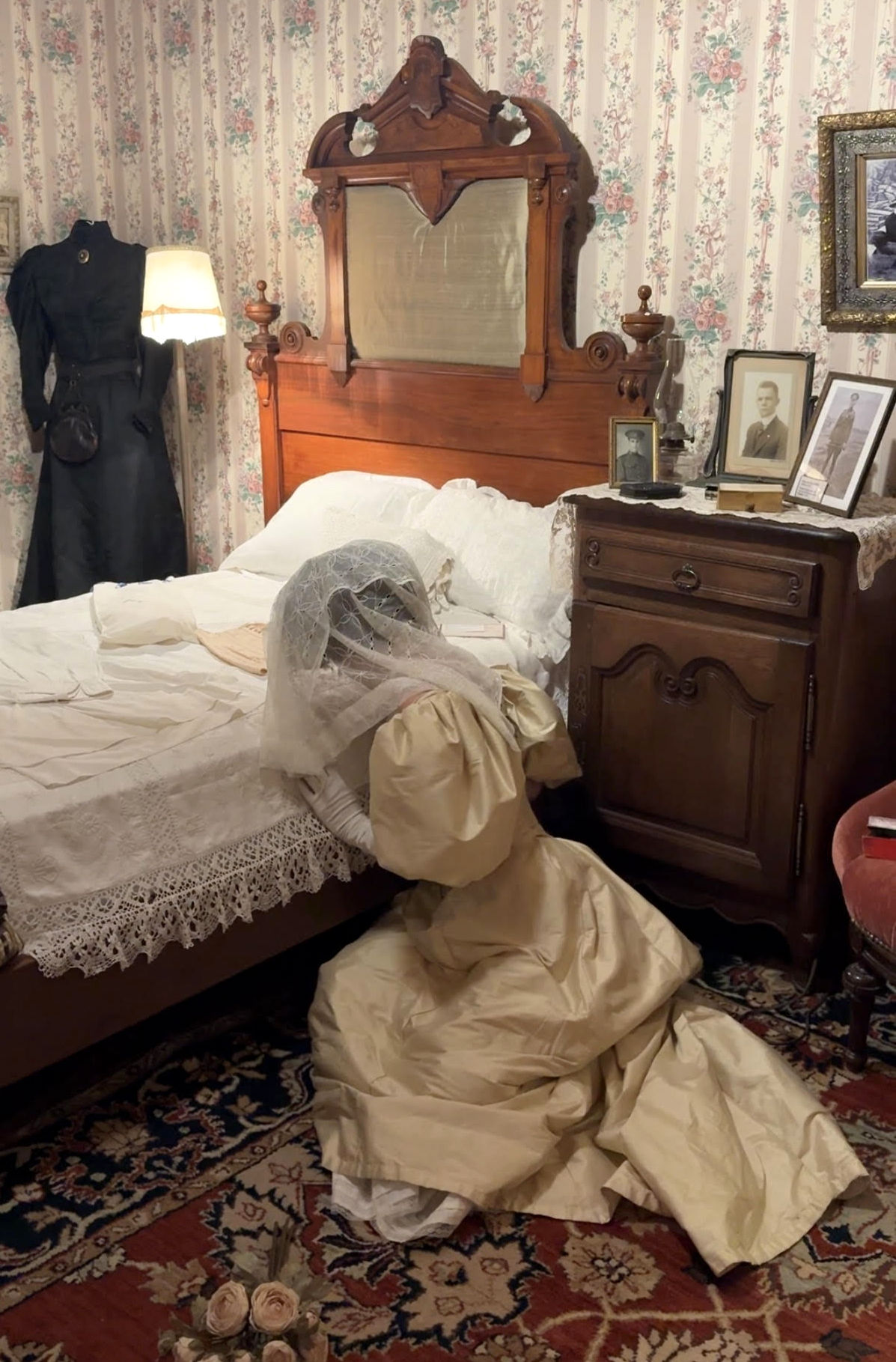 An Edwardian Bride wearing a silk wedding gown and veil sits crumpled on the floor alongside a bed. Her face is buried in the pillows.