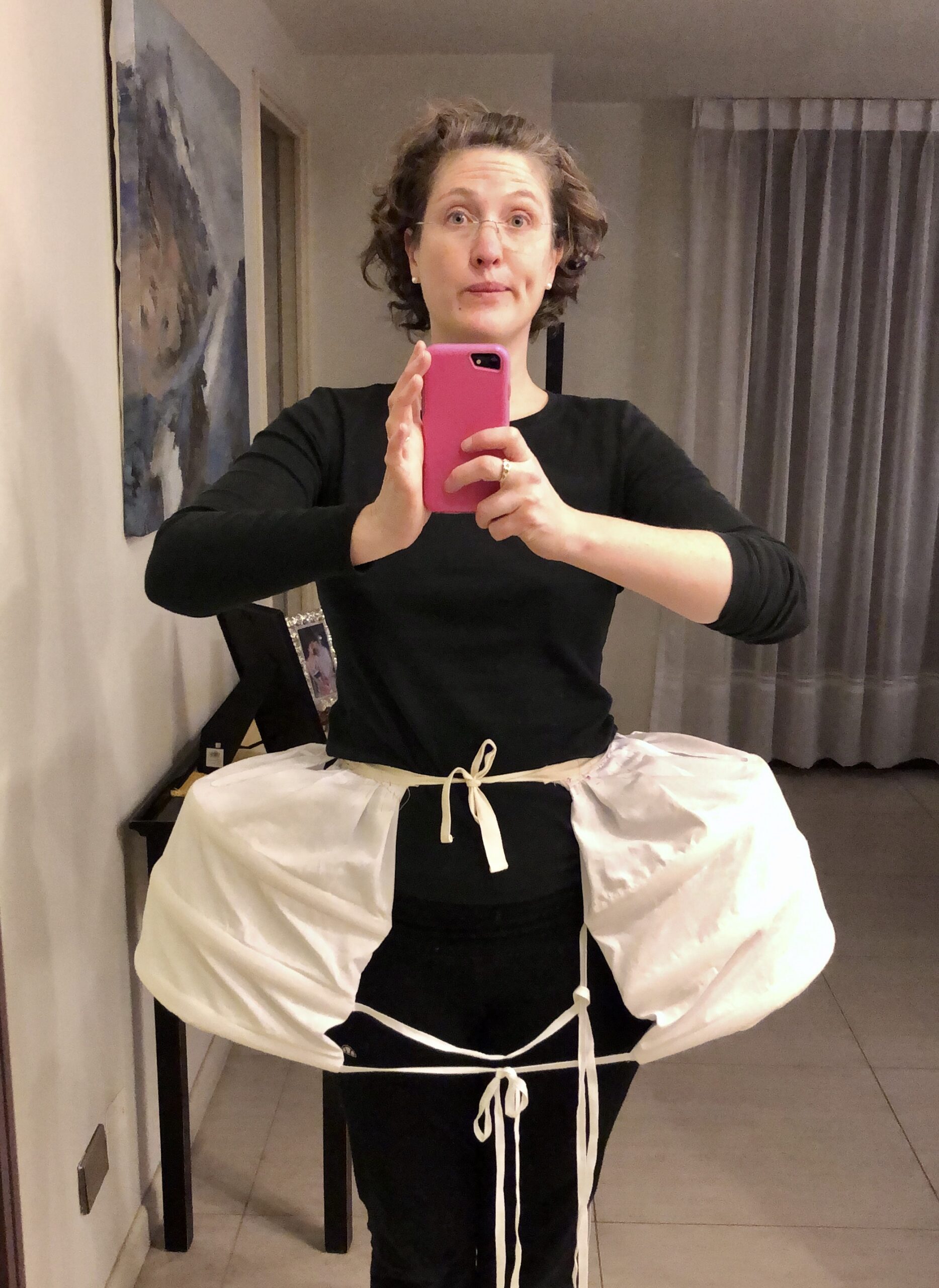 A woman faces the camera - she is wearing a pair of white 18th century pocket hoops over a black tshirt and leggings