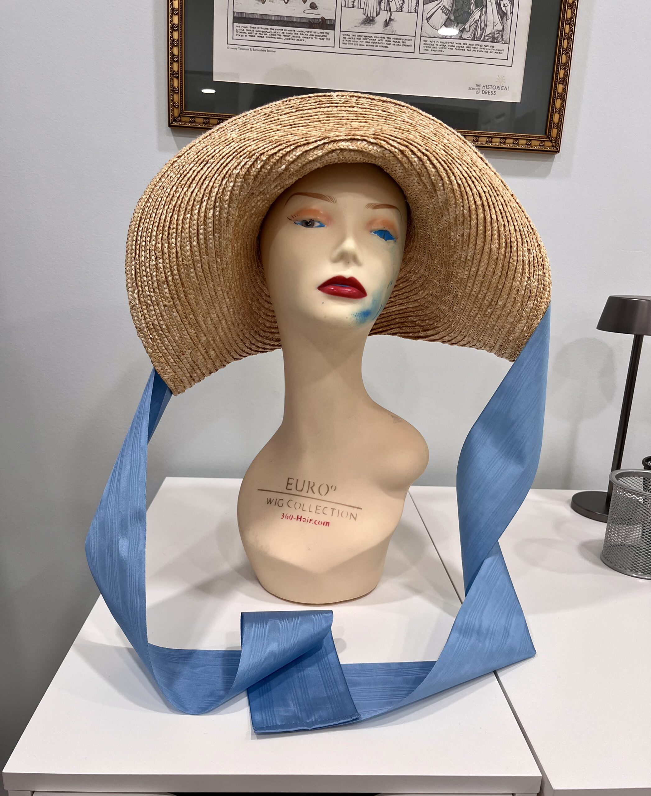 Front view of a high-crowned straw hat mounted on a wig mannequin. Blue moire ribbons stream down at each side
