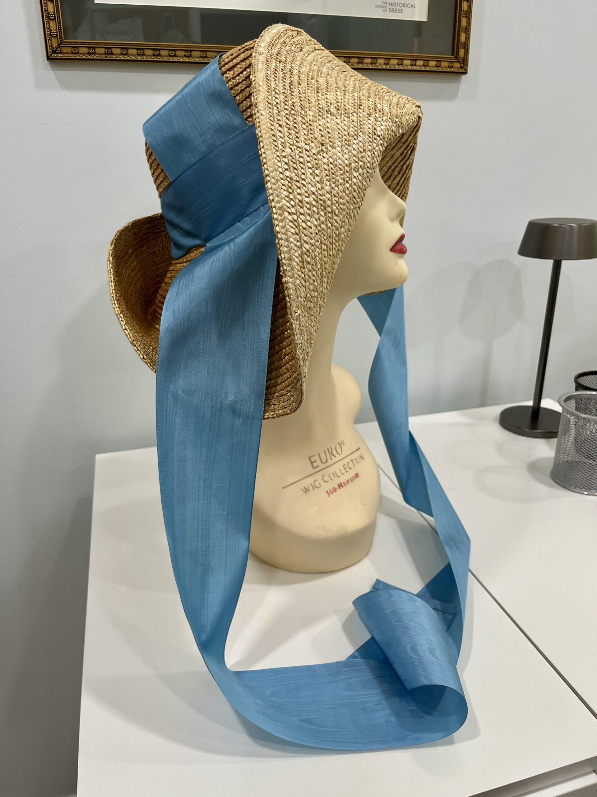 side view of a high-crowned straw hat mounted on a wig mannequin. Blue moire ribbons stream down at each side