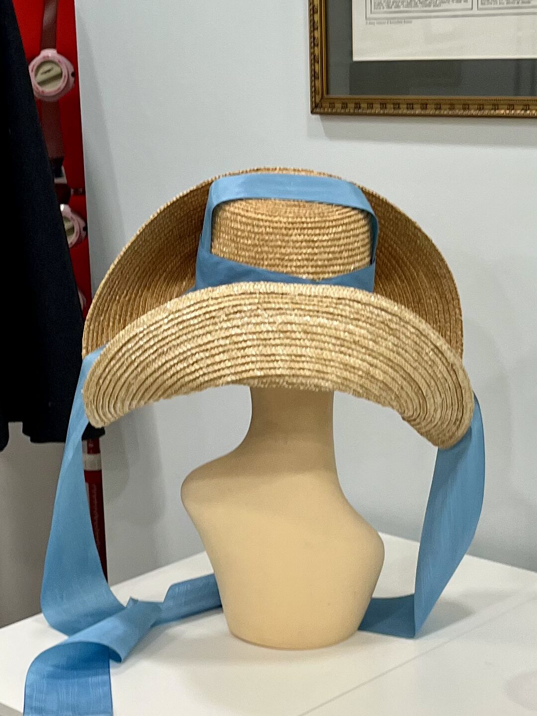 back view of a high-crowned straw hat mounted on a wig mannequin. Blue moire ribbons stream down at each side