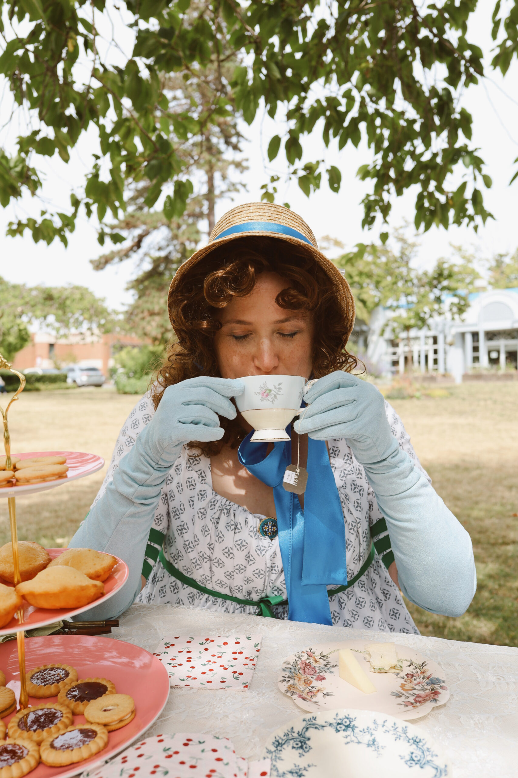 A woman in a straw bonnet and blue gloves drinks from a gold rimmed teapot at a white-draped table in a park