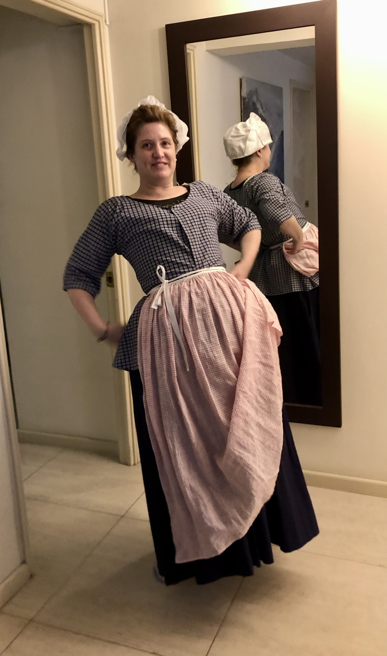 Tabubilgirl leans backwards. She is wearing a blue check shortgown, a long blue petticoat and a red check apron.