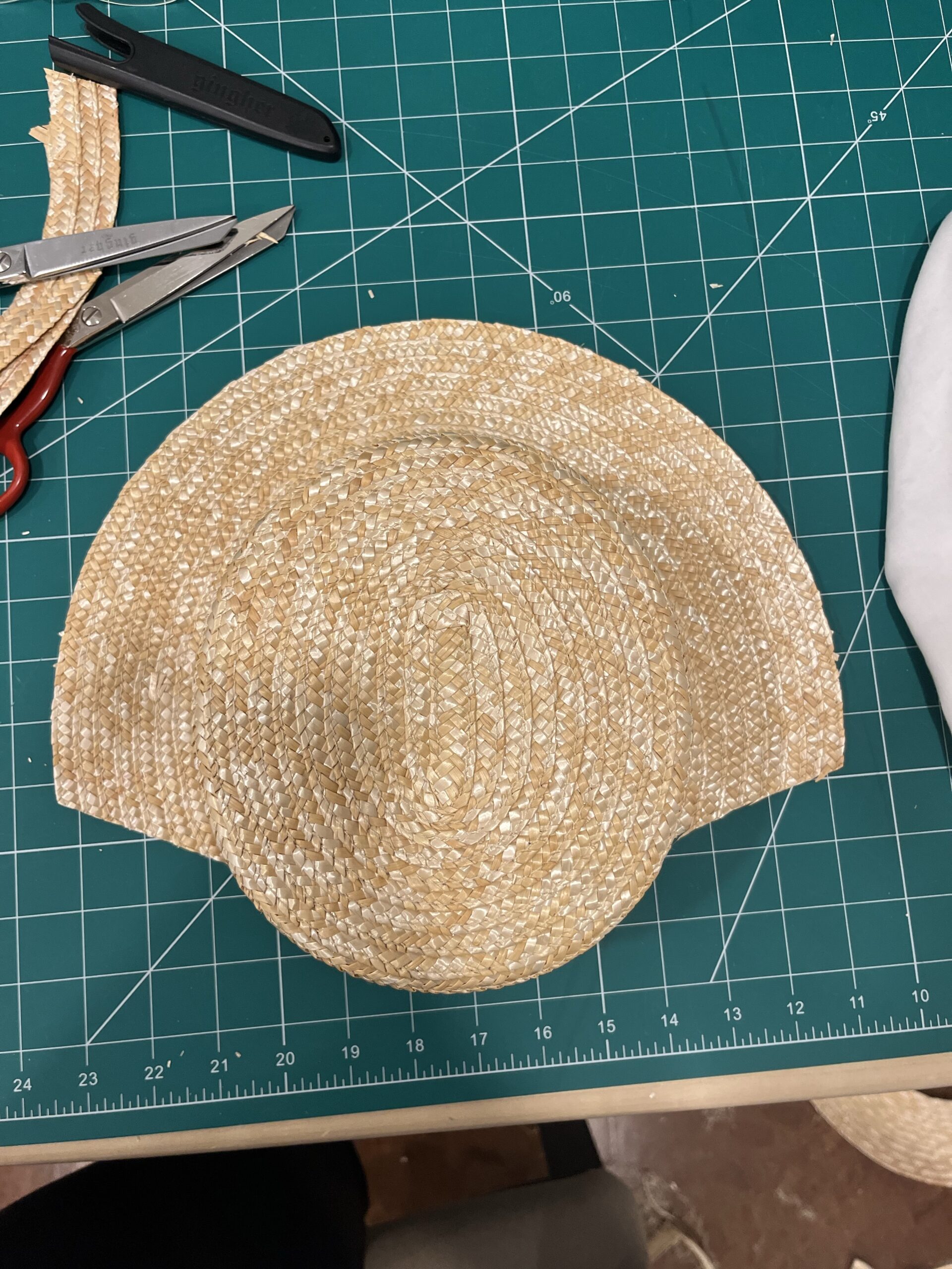 A straw hat sits on a green cutting board. Experiemental cuts in the brim have been made and a large chunk has been removed.