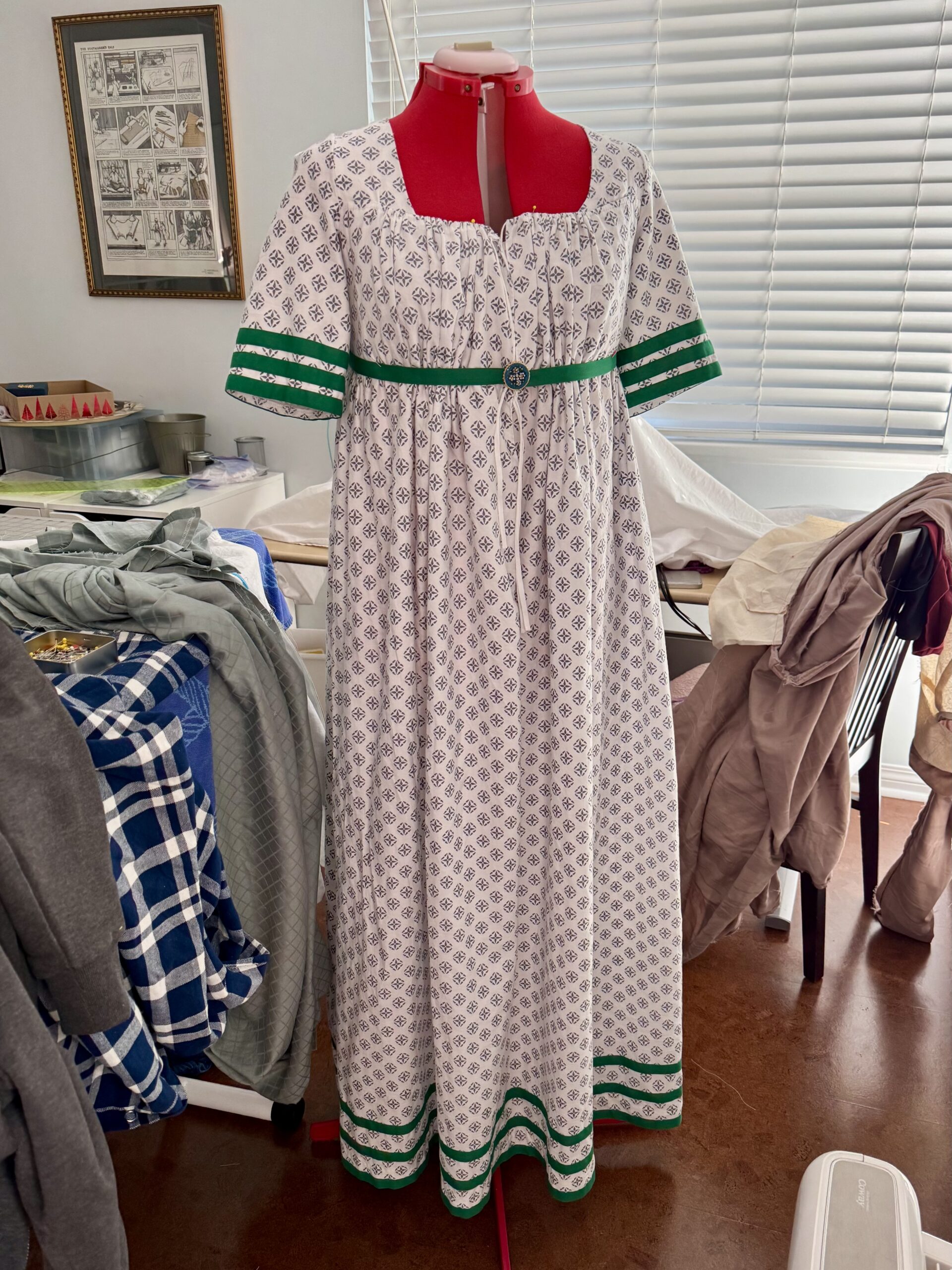 a 1790s dress sewn of white cotton printed with grey block print, and trimmed in three rows of racing stripes in green cotton tape on the hem and the sleeves. A green sash is pinned around the waist.