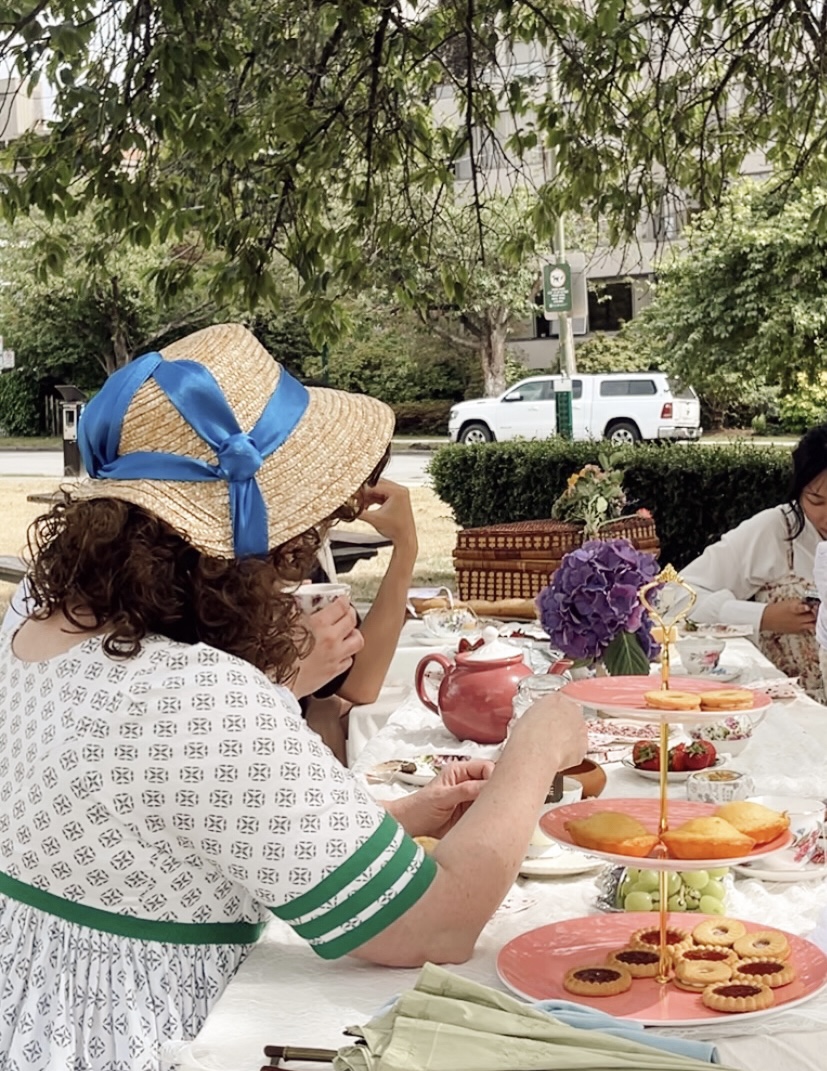 tabubilgirl sits at a picnic table wearing a block print 1798 round gown with green stripes on the sleeves- she is viewed from the back, with a very good view of the back of her 1809 straw bonnet