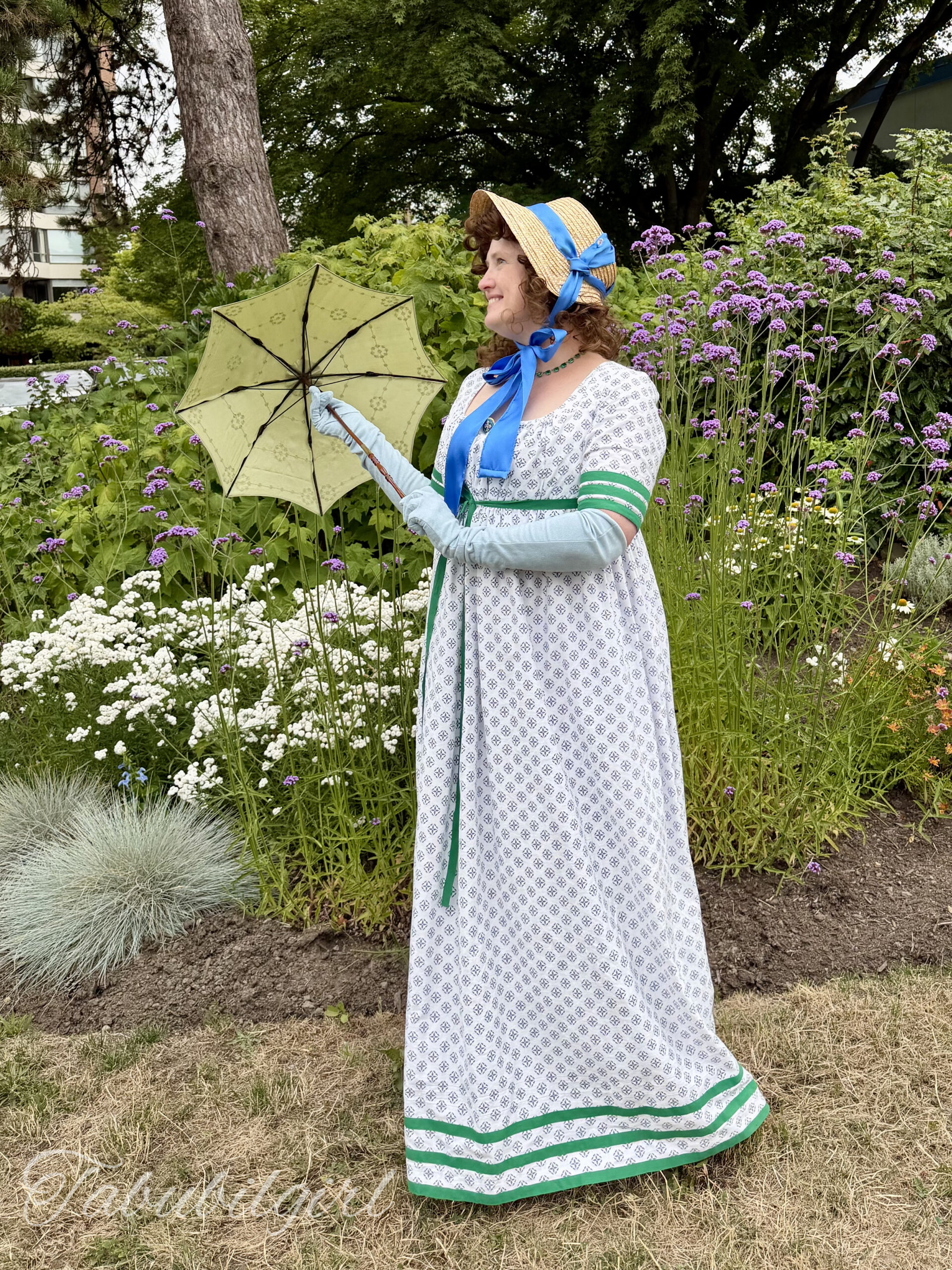 a woman stands in 3/4 position. She is wearing a 1798 round gown with green regency stripes, long blue gloves and a small straw bonnet. She is holding a tiny green silk parasol.