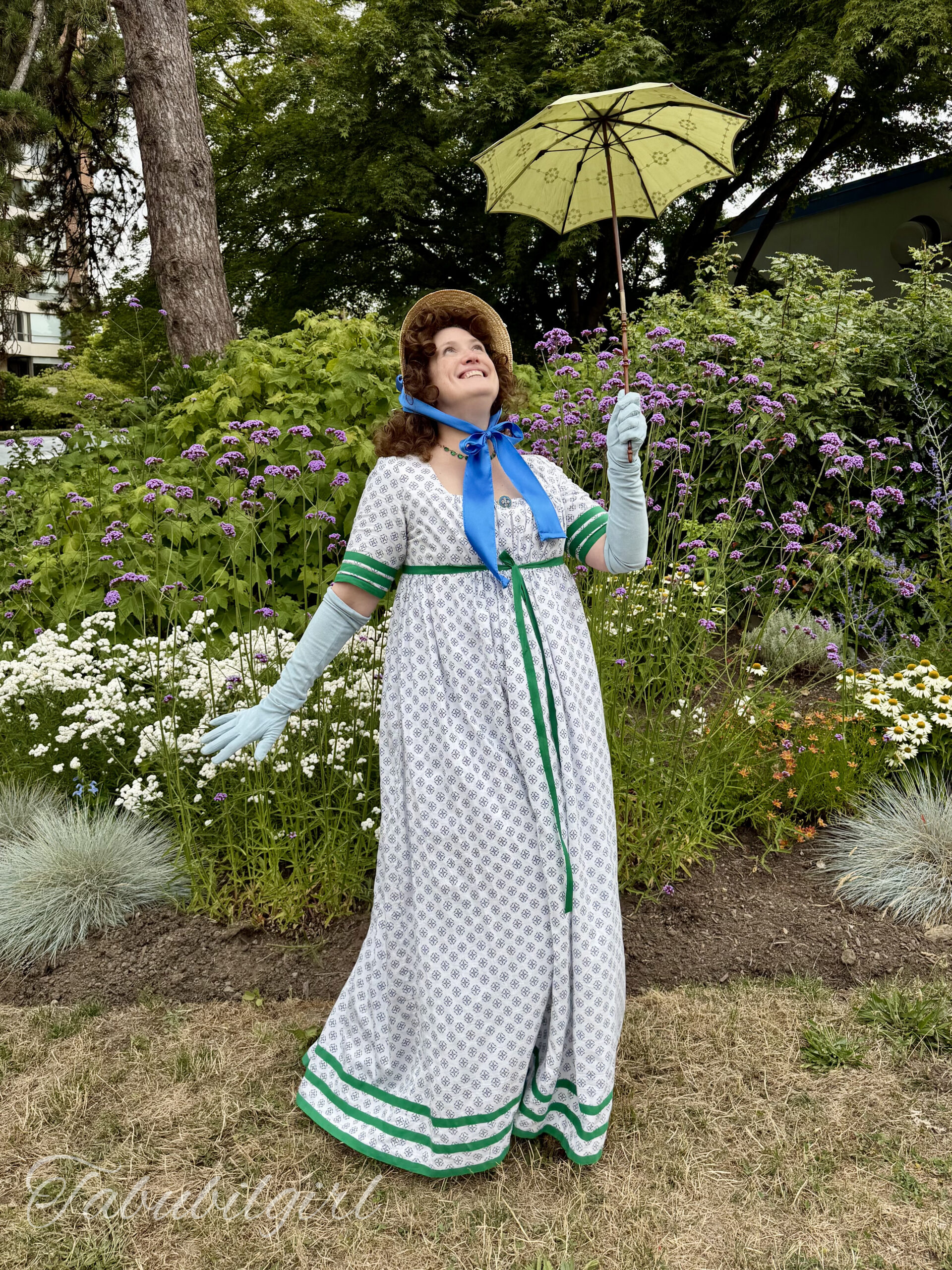 a woman stands in 3/4 position. She is wearing a 1798 round gown with green regency stripes, long blue gloves and a small straw bonnet. She is smiling and holding a tiny green silk parasol.
