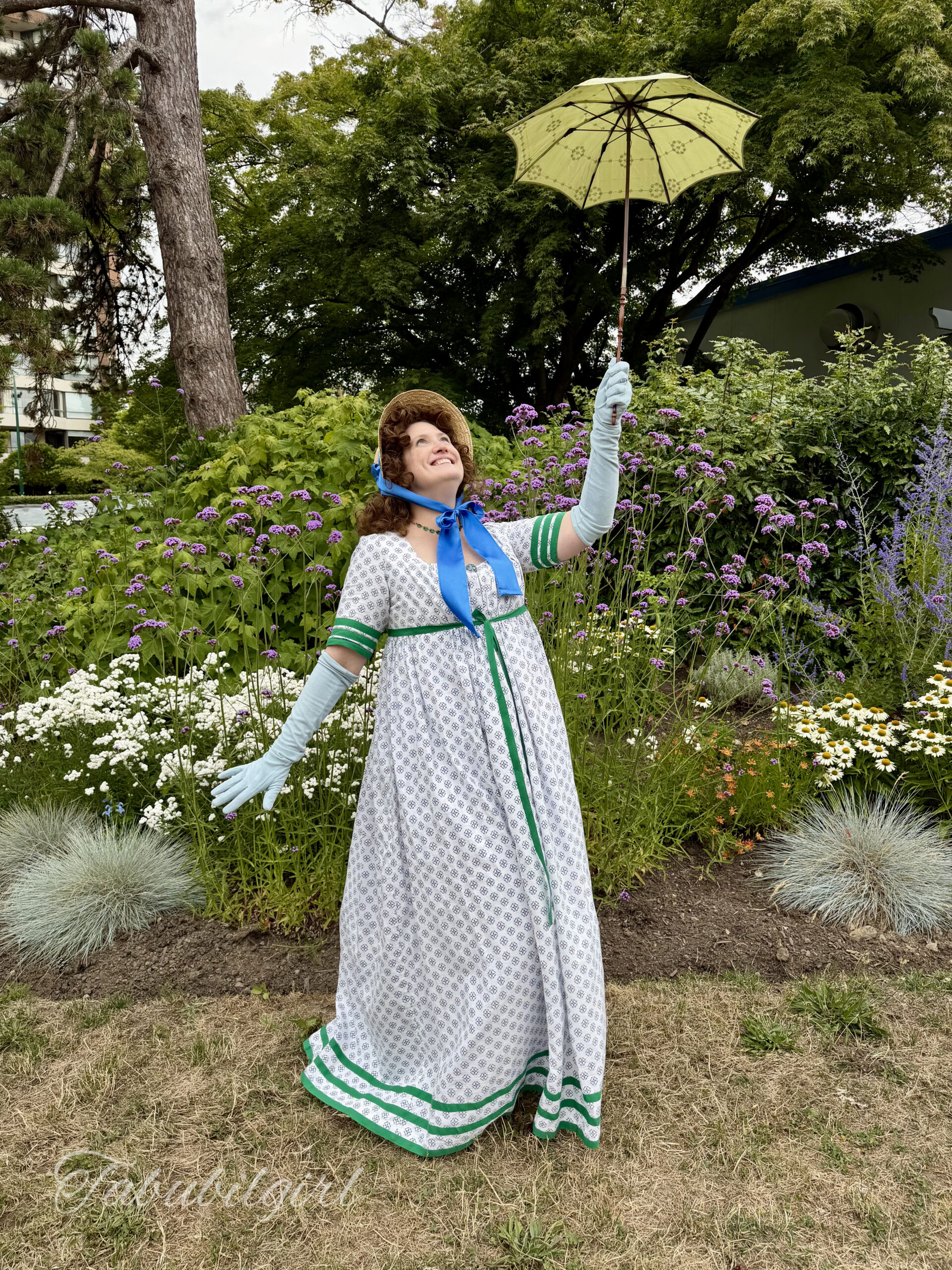 Tabubilgirl wears a 1790s round gown with rows of green racing stripes.  She is accessorized with a small straw bonnet and a pair of long blue gloves. She is smiling and raising a tiny green parasol high in the air.
