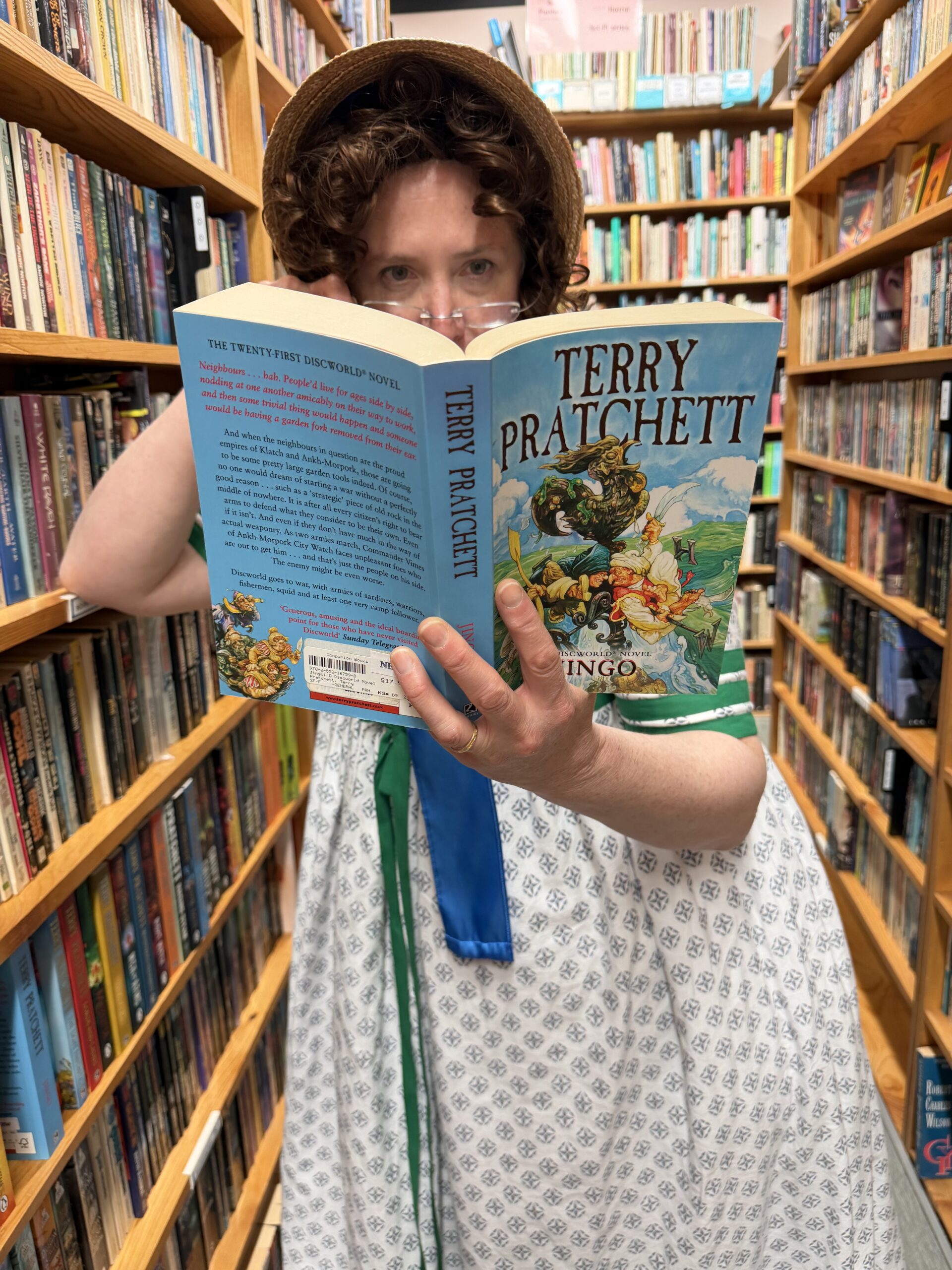 a woman in a block print regency round gown and straw bonnet holds a terry pratchett book up to the viewer