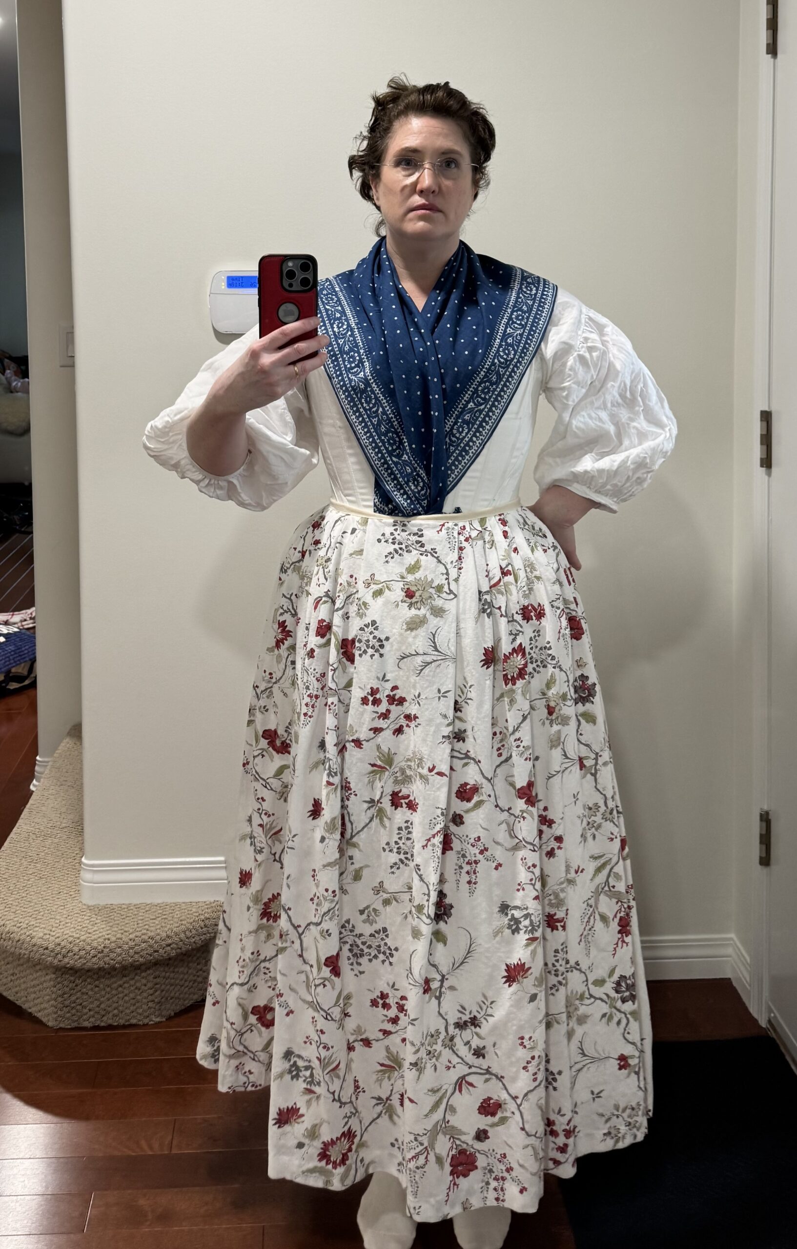 A woman faces the camera wearing an 18th century shift and stays below a cream chintz petticoat and indigo print kerchief.