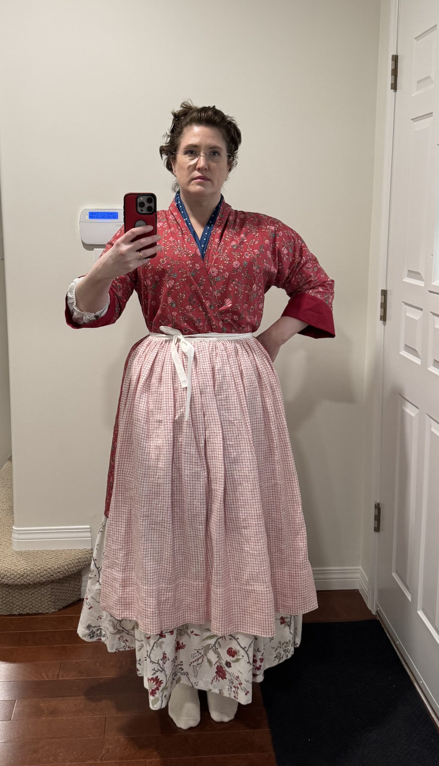 A woman faces the camera wearing a tomato red chinz bedgown, a red and white check apron and a cream chintz petticoat.